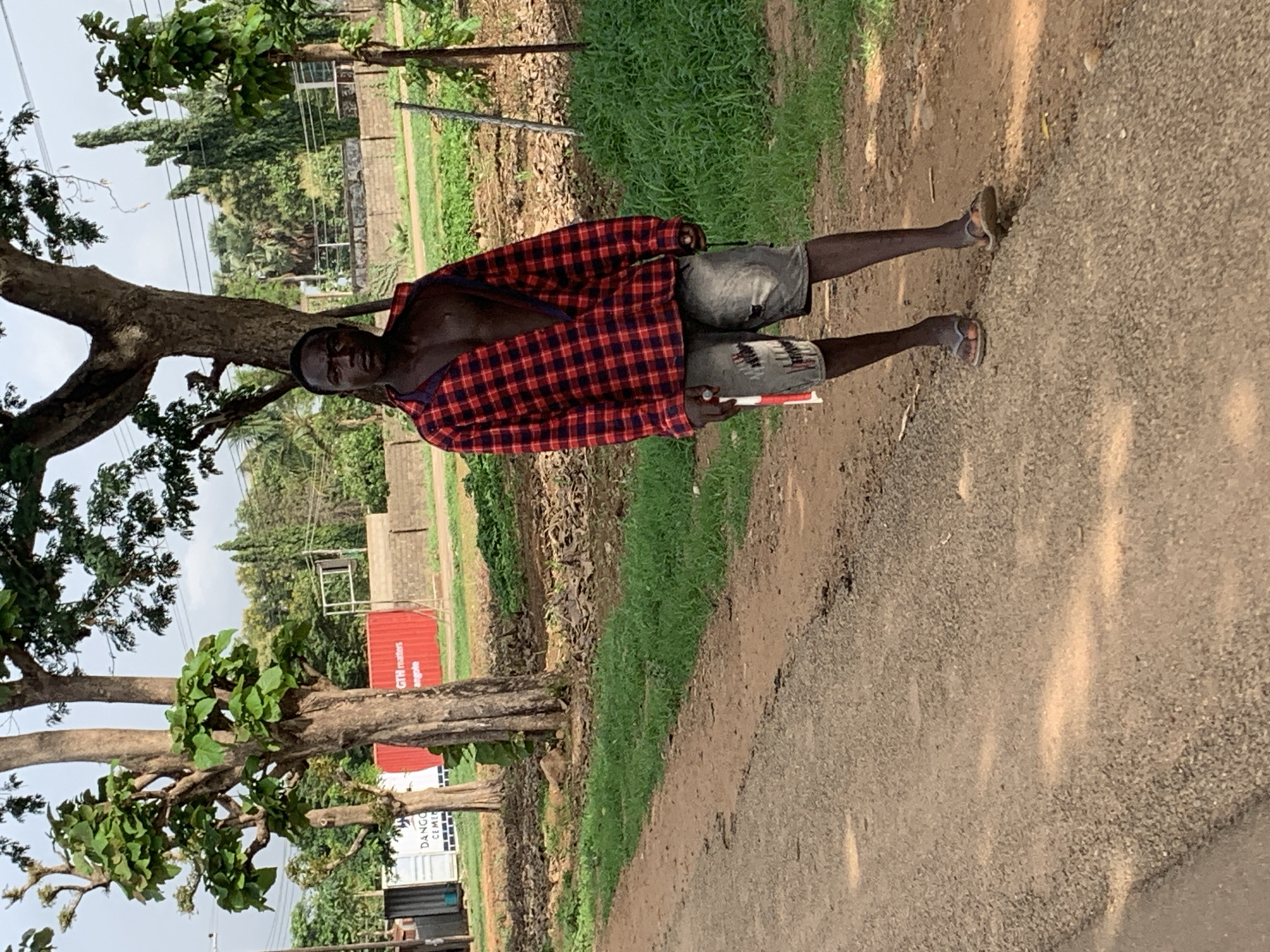 A person is standing on a gravel-covered path next to a tree in Agban, Nigeria. The person is wearing a red and black plaid shirt, gray shorts, and flip-flops. They are holding a white and red object in their hand. The background includes green grass, trees, and a red shipping container labeled "GTH Matters Angola." Another white shipping container labeled “DANGO CEMET” is also visible. Overhead electric lines can be seen in the distance.
