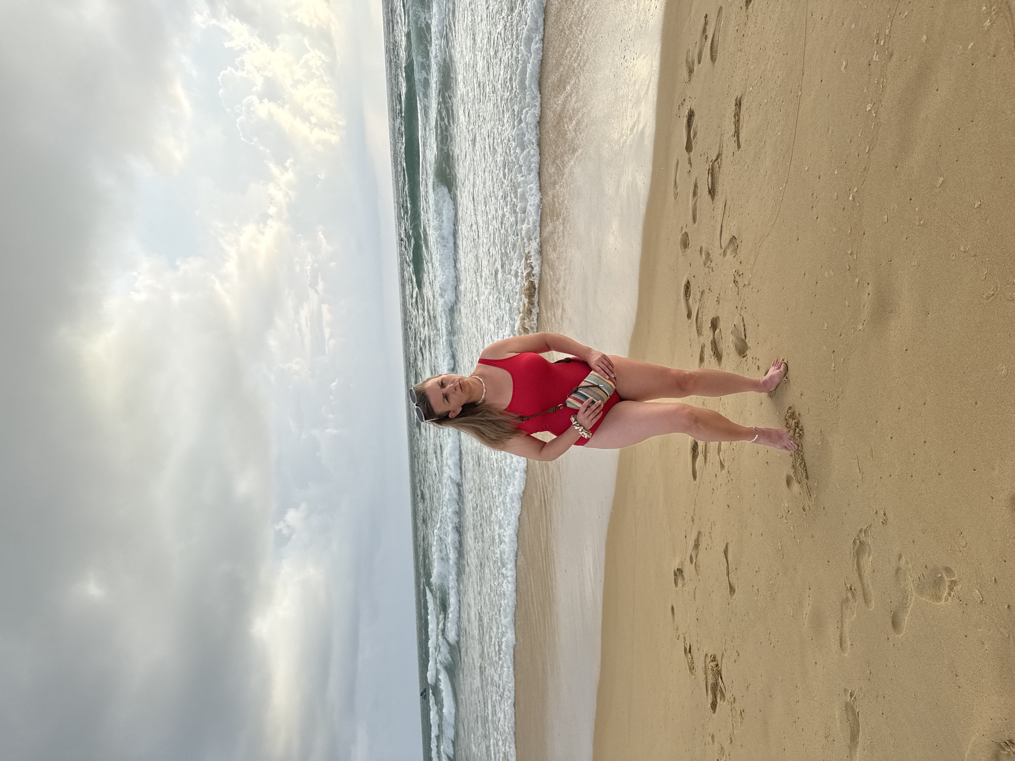 A woman in a red one-piece swimsuit stands on a sandy beach, holding a striped purse. She is facing forward and looking slightly to her left. The ocean with moderate waves is behind her, and the sky is overcast with dramatic clouds. Footprints are visible in the sand around her. The scene is located in Karon, Thailand.