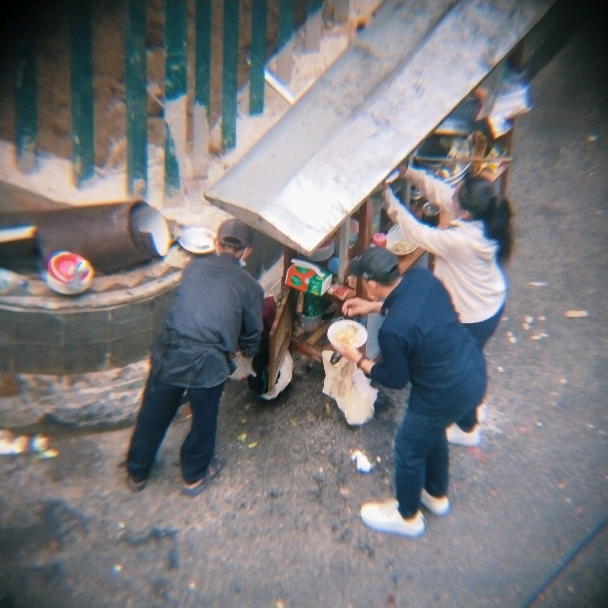 In Surabaya, Indonesia, a scene unfolds around a food stall. Three individuals are gathered. One person, wearing a dark shirt and dark pants, stands with their back to the camera, facing a stall laden with various items. Another person, in a dark blue shirt and dark pants, holds a white bowl containing food and appears to be serving or preparing it. They are wearing a dark baseball cap. A third person, in a light-colored hooded jacket, is leaning over the stall, reaching for something. The stall itself is a makeshift structure with a metal roof and wooden supports. Scattered around the stall are food items, containers, and what appears to be trash. The ground is paved with asphalt and pebbles. In the background, a green metal fence with vertical slats is visible. A large, dark cylindrical object is also present near the stall. The overall impression is of a busy street food vendor, possibly operating in a less-than-pristine environment.