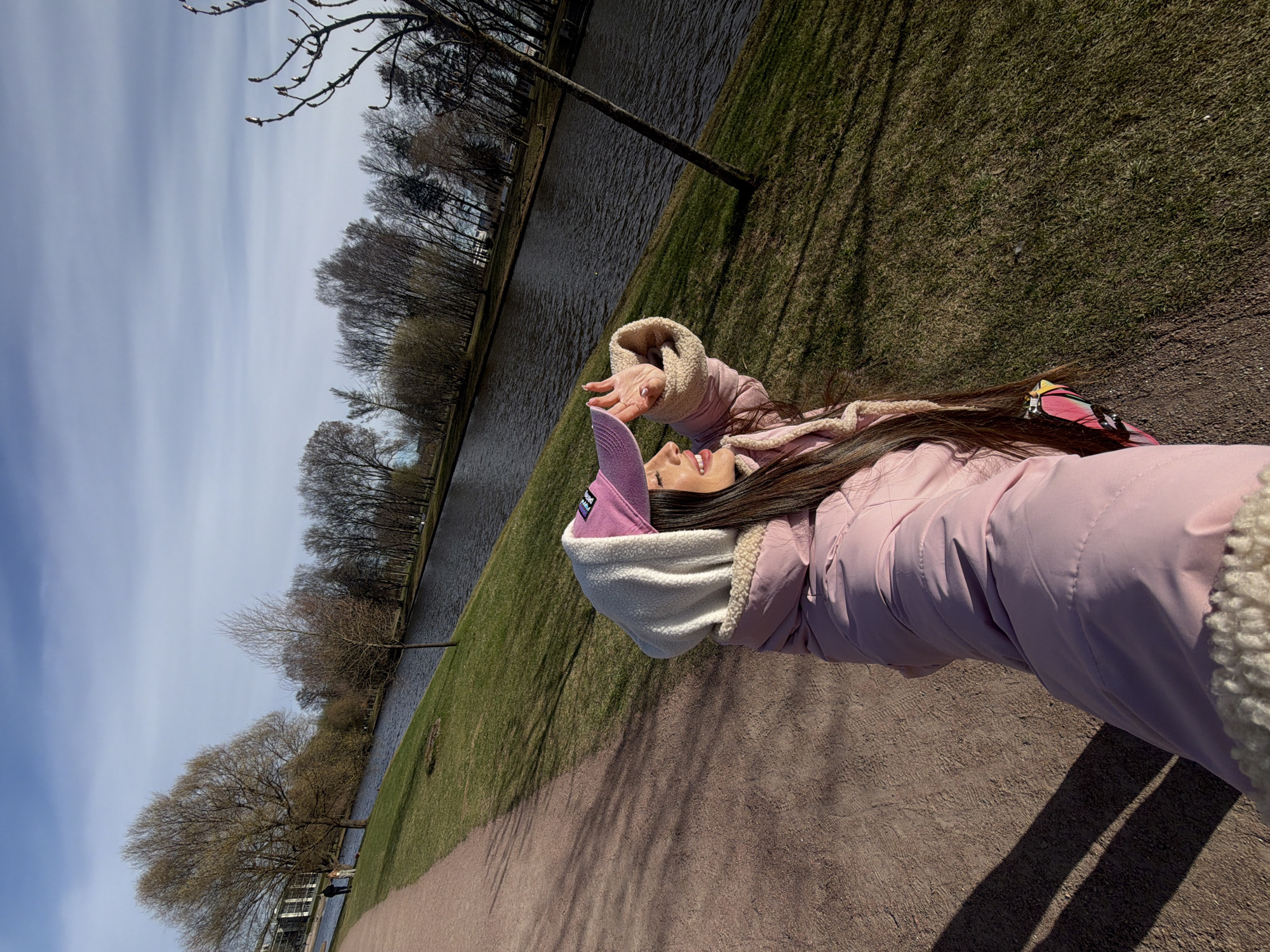 A young woman is the subject of this angled, outdoor photograph. She is lying on her back, looking up and toward the left with her arm raised in the air. Her hair is long, dark, and straight. She wears a pink jacket with a cream-colored lining, a white and pink hat, and sunglasses on top of her head.
The environment consists of a walkway, grass, a body of water, and trees. The scene appears to be in a park during the early spring, as the trees lack leaves. The location is in the city of Saint Petersburg, Russia.