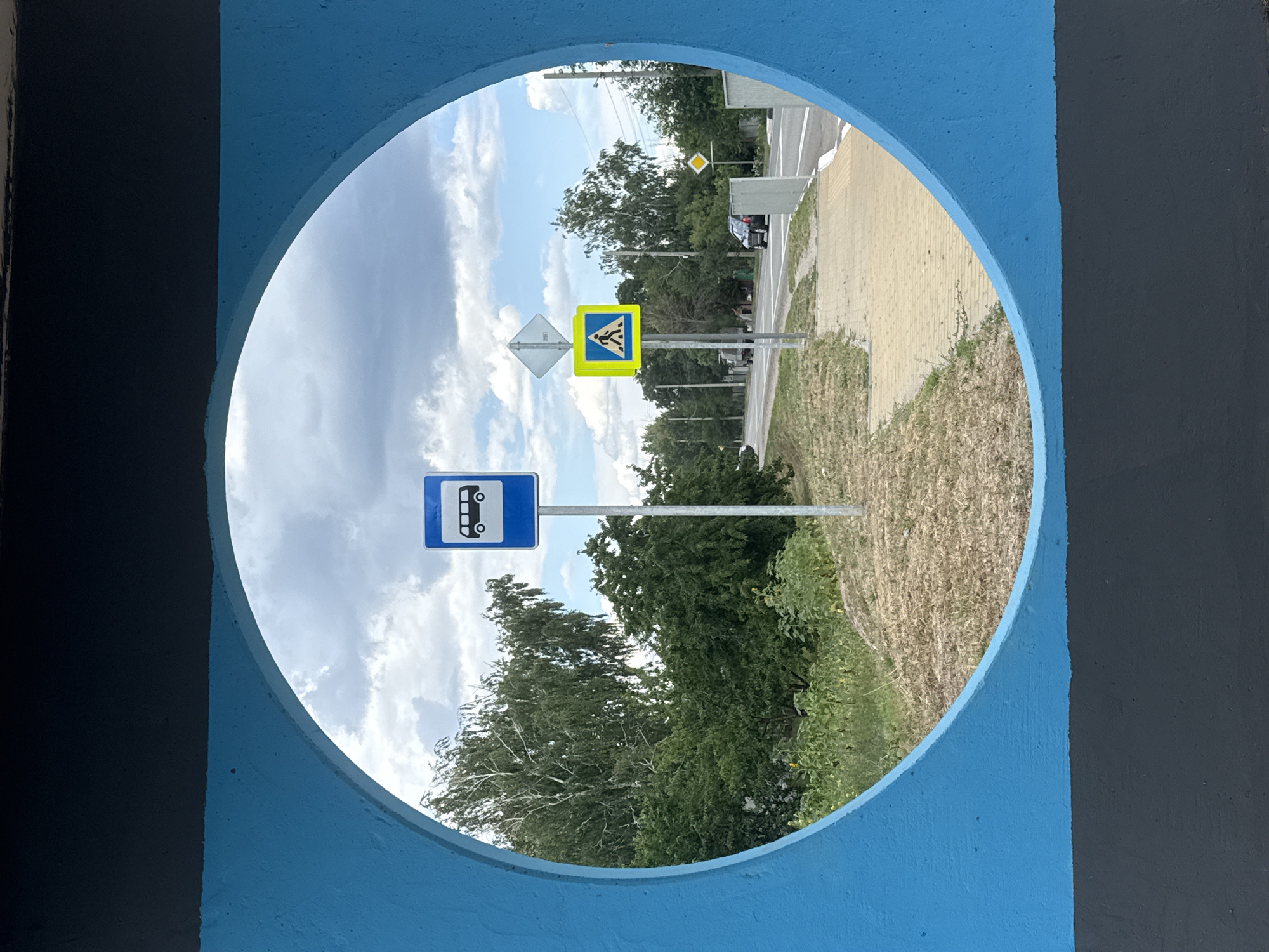 The image, framed by a circular opening in a blue and black structure, captures a view of a street scene in Shulhynka, Ukraine. On the right side of the view, a paved sidewalk runs parallel to a road. Across the road, a grassy area and trees are visible. A black car is parked on the right side of the road. On the left side of the road, a bus stop sign and a pedestrian crossing sign are prominently displayed. The sky above is filled with clouds. The foreground is occupied by the blue and black painted structure, which forms the circular frame. The overall impression is that of a sunny day with a clear view of urban infrastructure and surrounding greenery.