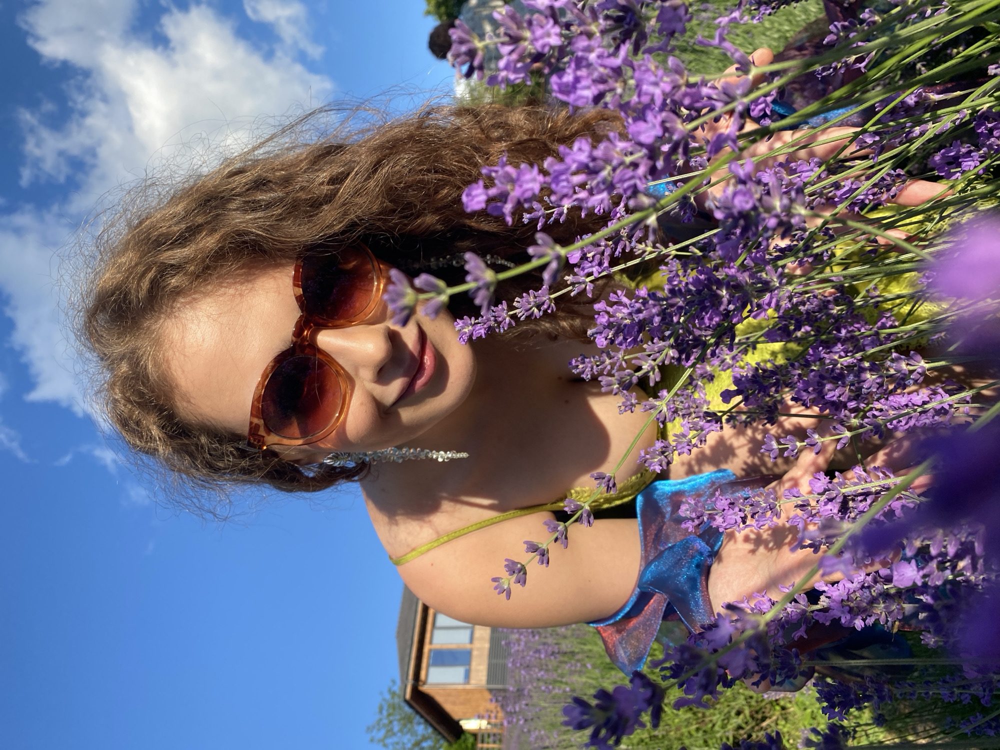 The image depicts a young woman with curly brown hair, wearing sunglasses and a yellow and blue dress, crouched in a lavender field. She is looking at the camera and smiling. Her hands are reaching out towards the lavender flowers. In the background, there is a wooden house and a clear blue sky with some clouds. The location is identified as Verkhneye Uchdere, Russia. The overall scene suggests a warm, sunny day in a rural setting.