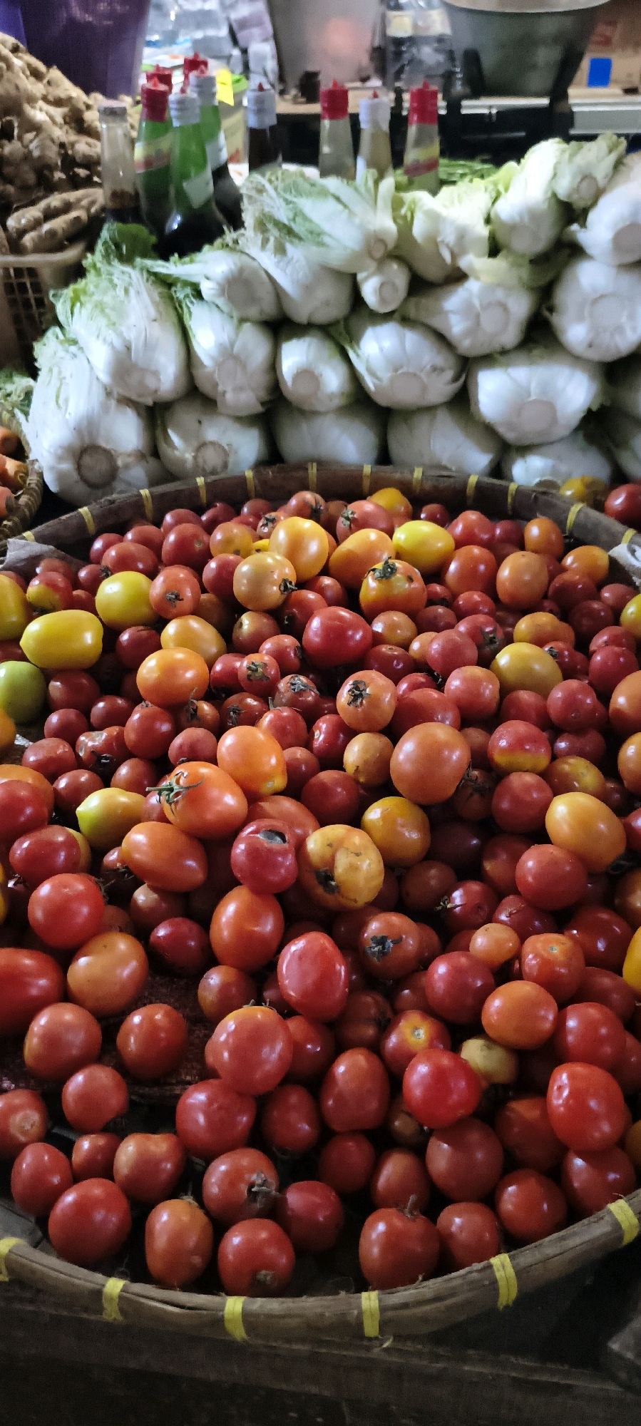This image depicts a market stall in Weru Lor, Indonesia, filled with a vibrant display of fresh produce. In the foreground, a woven basket overflows with a multitude of tomatoes, varying in size, shape, and color. They range from deep reds and bright oranges to sunny yellows and even a hint of green, with some showing slight imperfections characteristic of fresh, unpackaged produce. Behind the tomatoes, several heads of napa cabbage are stacked, their pale green and white leaves creating a textured backdrop. In the background, a pile of ginger root is visible in a basket to the left, while several bottles with red and white caps, possibly condiments or sauces, are lined up to the right, suggesting the diverse offerings of the market. The lighting is natural, characteristic of an outdoor or semi-open market setting. The overall scene conveys a sense of abundance and the lively atmosphere of a local Indonesian marketplace.