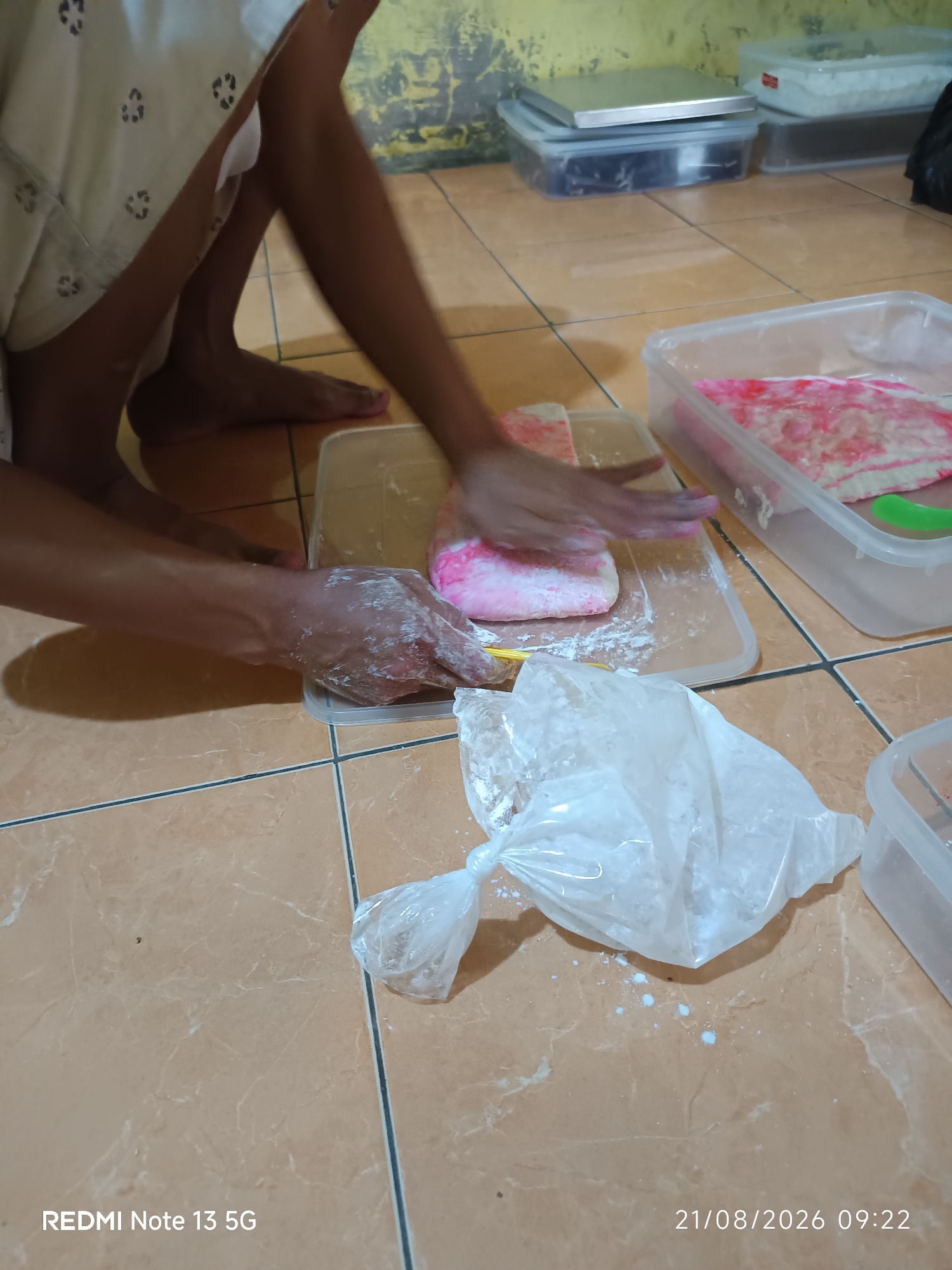 In an indoor setting with a tiled floor, a person is engaged in food preparation. Their hands, dusted with white powder, are actively working with a pink and white striped dough-like substance placed on a clear plastic tray. Adjacent to this tray is a larger clear plastic container holding a similar pink and white striped material. To the right, another clear plastic container is visible, partially obscured. In the foreground, a translucent plastic bag filled with white powder lies on the floor, with some powder spilled around it. The background shows a yellow-grey wall and several stacked plastic containers. The image appears to be taken in the city of Talaga Sari, Indonesia, with a timestamp of 21/08/2026 09:22.