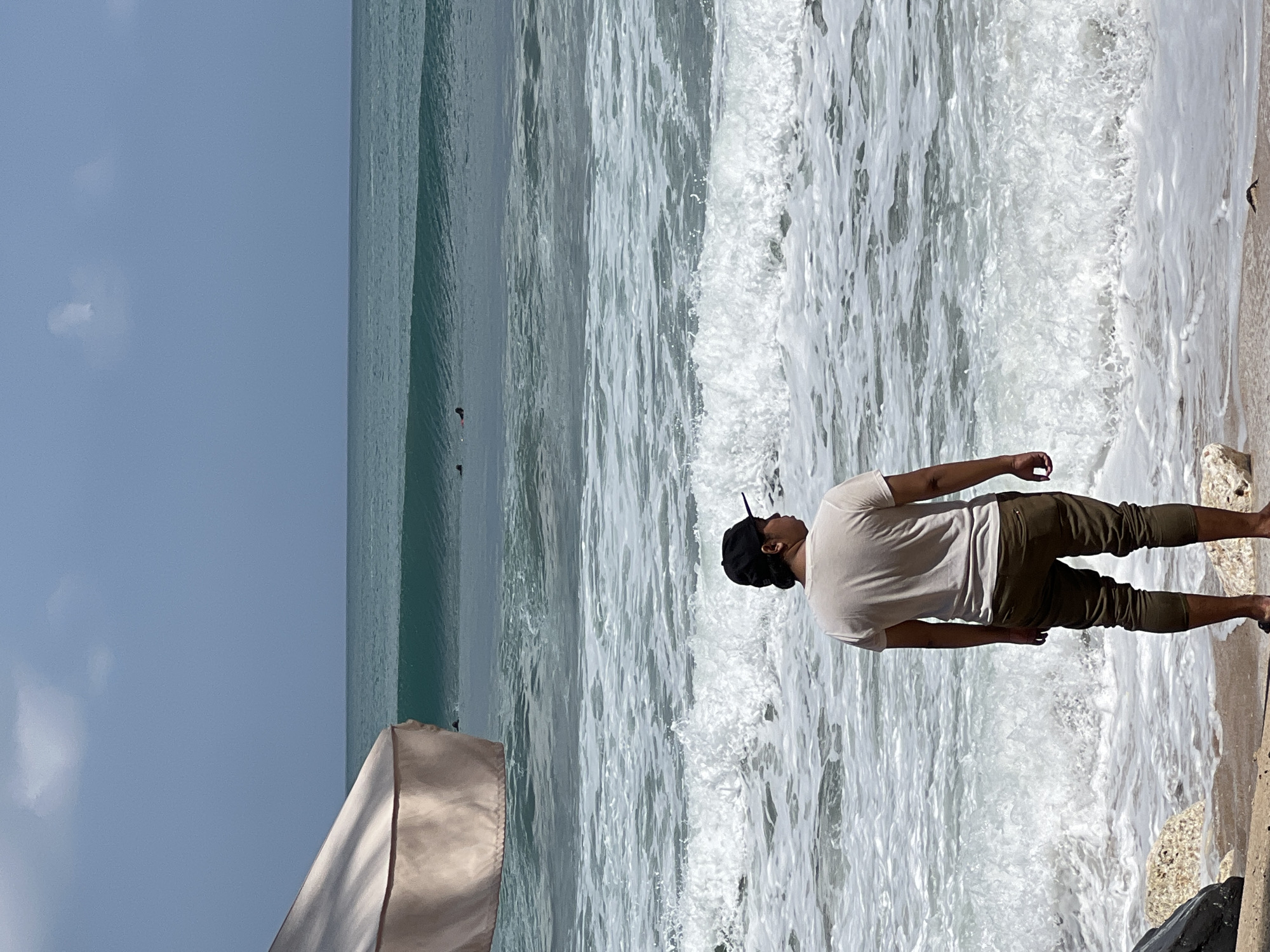 A person stands on a sandy beach in Kuta, Indonesia, facing the ocean. The individual is wearing a light-colored t-shirt and dark pants, with a black baseball cap. They are positioned with their back to the camera, looking out at the waves. The ocean is visible with a series of breaking waves and the distinct turquoise color of the water. In the distance, several surfers are in the water, some appearing to be waiting for waves or riding them. A portion of a light-colored beach umbrella is visible on the left side of the frame, casting a shadow. The sky is partly cloudy and a light blue. The foreground shows the sand and some rocks at the water's edge.
