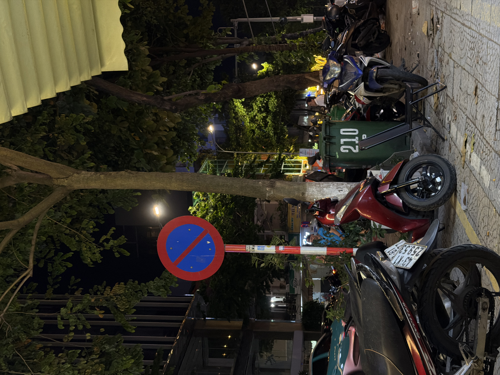 The image depicts a street scene at night, likely in Đà Nẵng, Vietnam. Several motorcycles are parked along a sidewalk with a textured, tiled surface. A large green trash bin with the number "210" visible on its side is also present. A "no parking" sign, featuring a red circle with a blue interior and a red diagonal line, stands on a pole with red and white stripes.
The surrounding environment includes dense foliage from trees and buildings in the background, with some streetlights illuminating the area. One motorcycle is a bright red scooter. Another is a blue and white motorcycle. The scene appears to be relatively calm, with no immediate indication of any specific event. The lighting suggests it is nighttime. A license plate with the text "59 S3 059.53" is visible on one of the motorcycles. Some debris is scattered on the sidewalk.