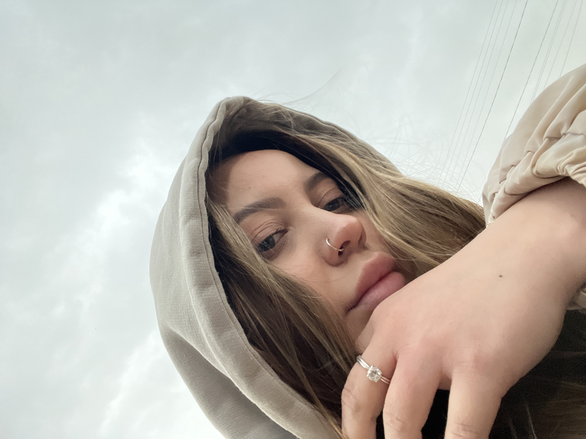 A close-up, low-angle shot shows a young woman wearing a hooded sweatshirt. The hood is up, partially framing her face. She has fair skin, light brown hair, and a nose ring. Her hand is near her face, and she wears a ring with a square-cut stone on her ring finger. The background is a cloudy sky, with power lines visible. The location in Novopokrovskaya, Russia, is not evident in the image.