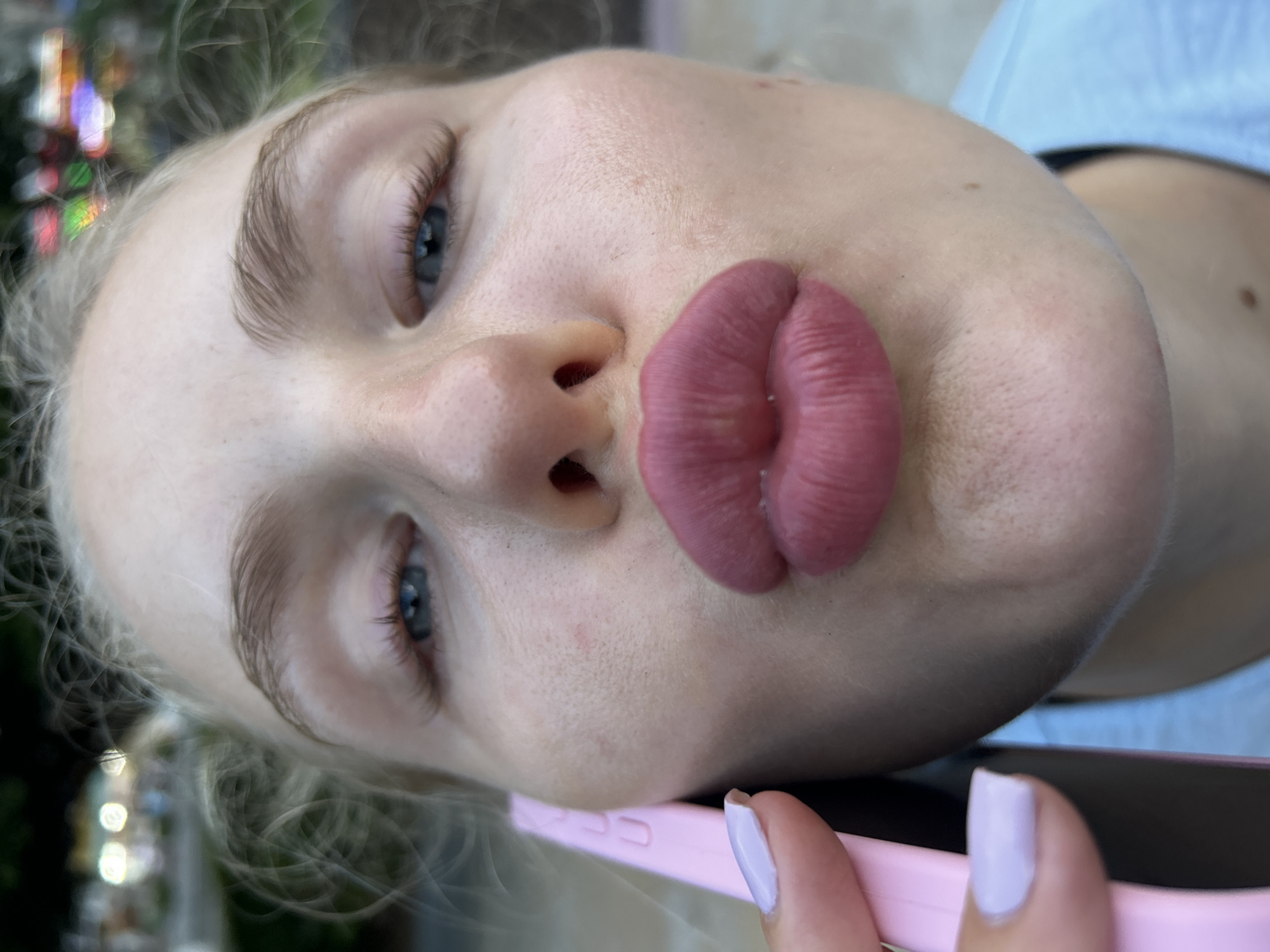 The image is a close-up, eye-level shot of a woman's face, as she is holding a pink phone to her right ear and looking directly into the camera with her lips puckered. Her blonde hair is pulled back, and her blue eyes are visible. The background is blurred, showing some out-of-focus lights and greenery, suggesting an outdoor setting, possibly a street or park in Nha Trang Ward, Vietnam. The woman is wearing a light blue shirt. Her fingernails are painted a pale lavender color.