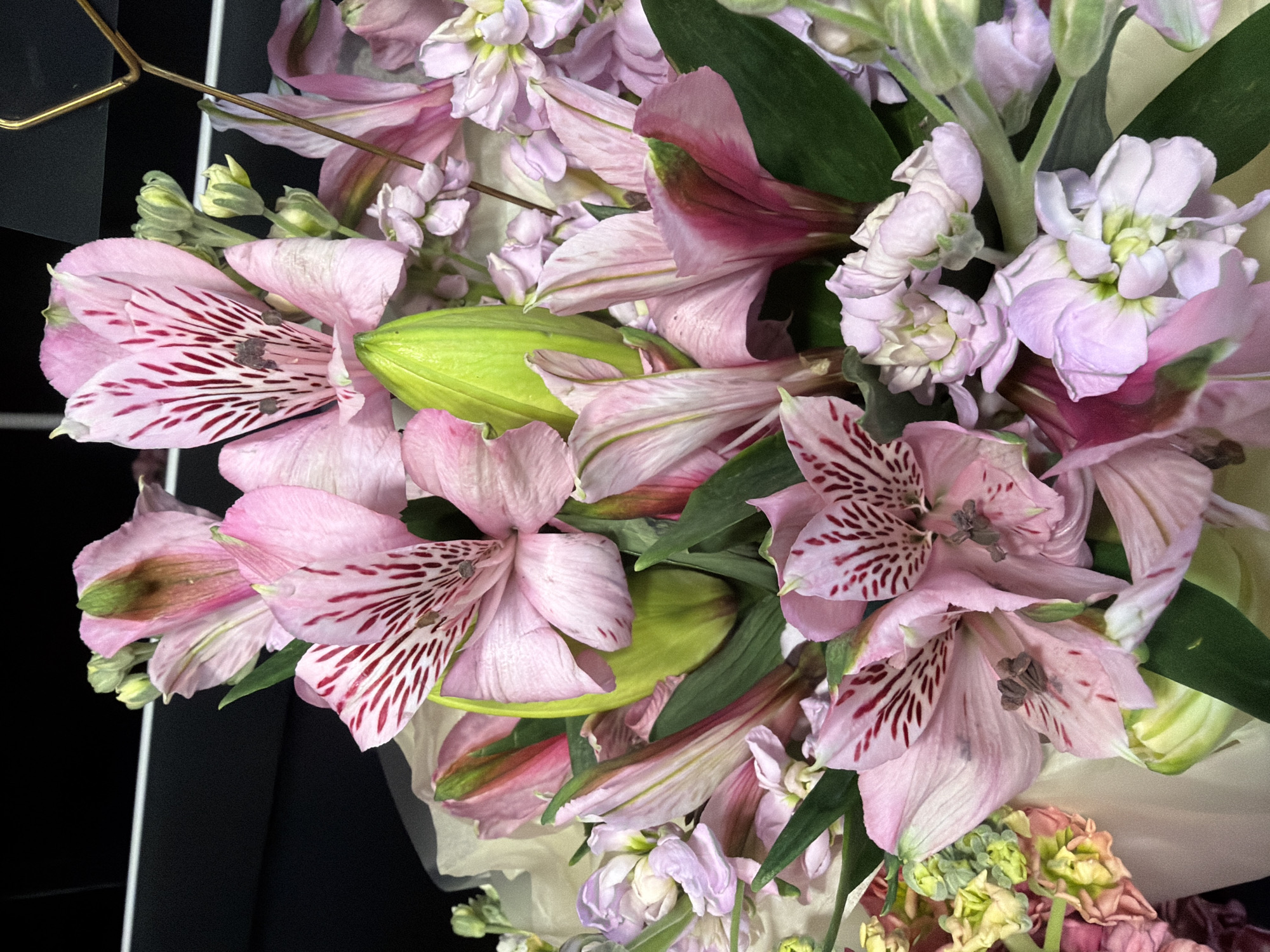 The image displays a close-up view of a bouquet of pink and lavender flowers, primarily alstroemeria and possibly stock flowers. The alstroemeria flowers are large and open, with delicate pink petals marked by deep red streaks radiating from the center. Many are adorned with green buds and leaves. Interspersed among them are clusters of smaller, pale lavender flowers, likely stock flowers, with multiple layers of tiny petals. The background is dark and out of focus, suggesting an indoor setting, possibly a florist shop or a home. The lighting is soft, highlighting the textures and colors of the blooms. There are no people or any identifiable urban or natural environmental features present in the image. The scene appears to be a still-life arrangement of flowers.