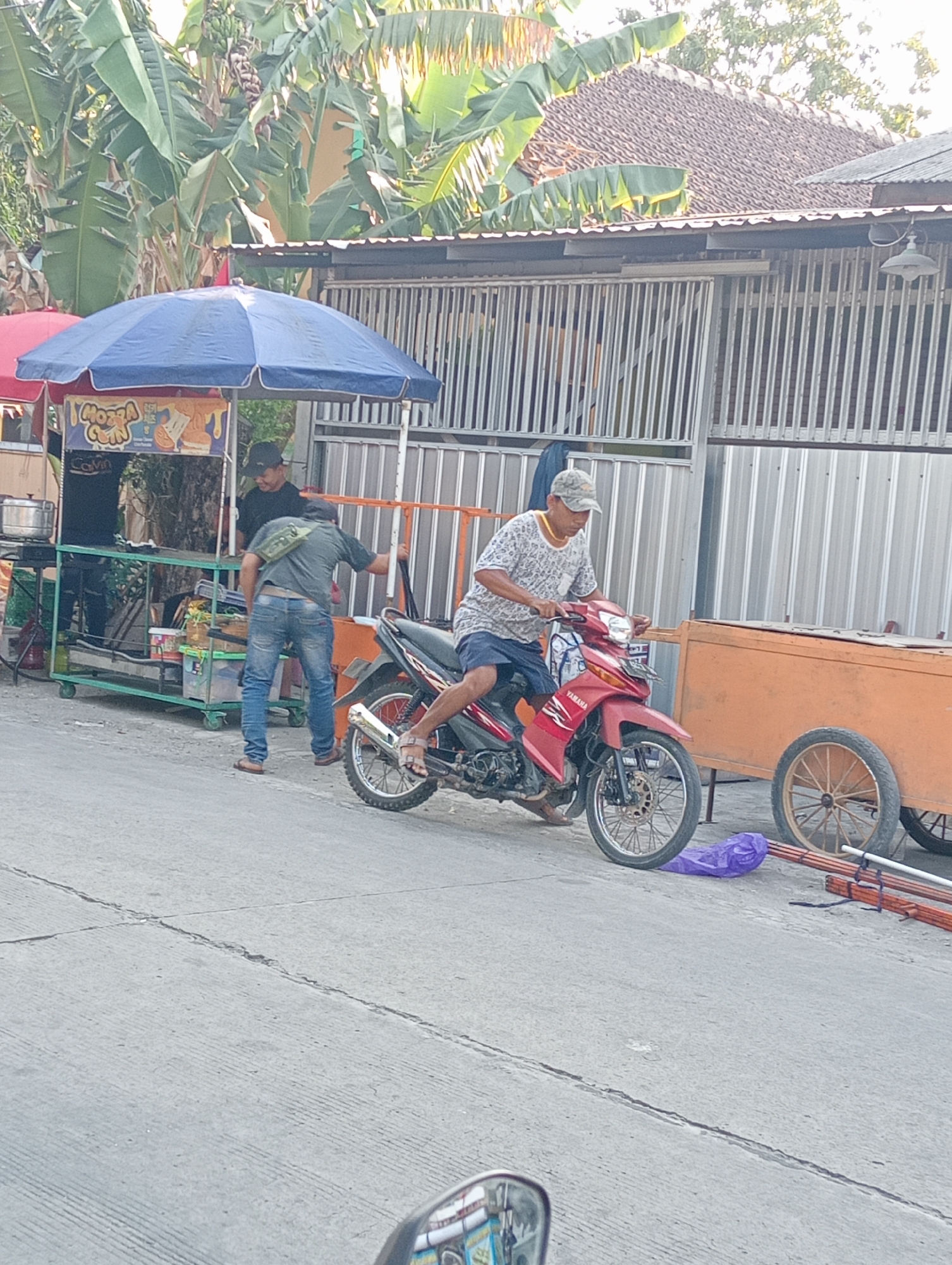 The image depicts an outdoor scene on a street in Tanjunganyar, Indonesia, with a focus on a small food stall and its surroundings. In the foreground, a red motorcycle is parked, with its rider on the left side of the frame. The rider, wearing a patterned t-shirt, shorts, and a cap, is seated on the motorcycle, facing towards the left. To the right of the motorcycle, a large, orange, two-wheeled cart is visible. A purple plastic bag and some wooden poles are on the ground near the cart.
Behind the motorcycle, a food stall with a blue canopy is set up. Two men are interacting near the stall. One man, wearing a black t-shirt and a cap, is standing behind the counter of the stall, looking towards the other man. The second man, dressed in a dark t-shirt and blue jeans, is standing in front of the stall, leaning slightly forward and facing away from the camera. He has a green bag slung across his shoulder. The stall itself appears to be selling food items, with some signage visible on the front.
The background shows a building with a corrugated metal fence or wall, and behind that, a tiled roof. Lush green banana trees are prominent in the upper portion of the image, suggesting a tropical or sub-tropical environment. The street surface is paved concrete, with visible cracks and lines. The overall lighting suggests it is daytime, likely late morning or early afternoon, given the shadows and brightness.