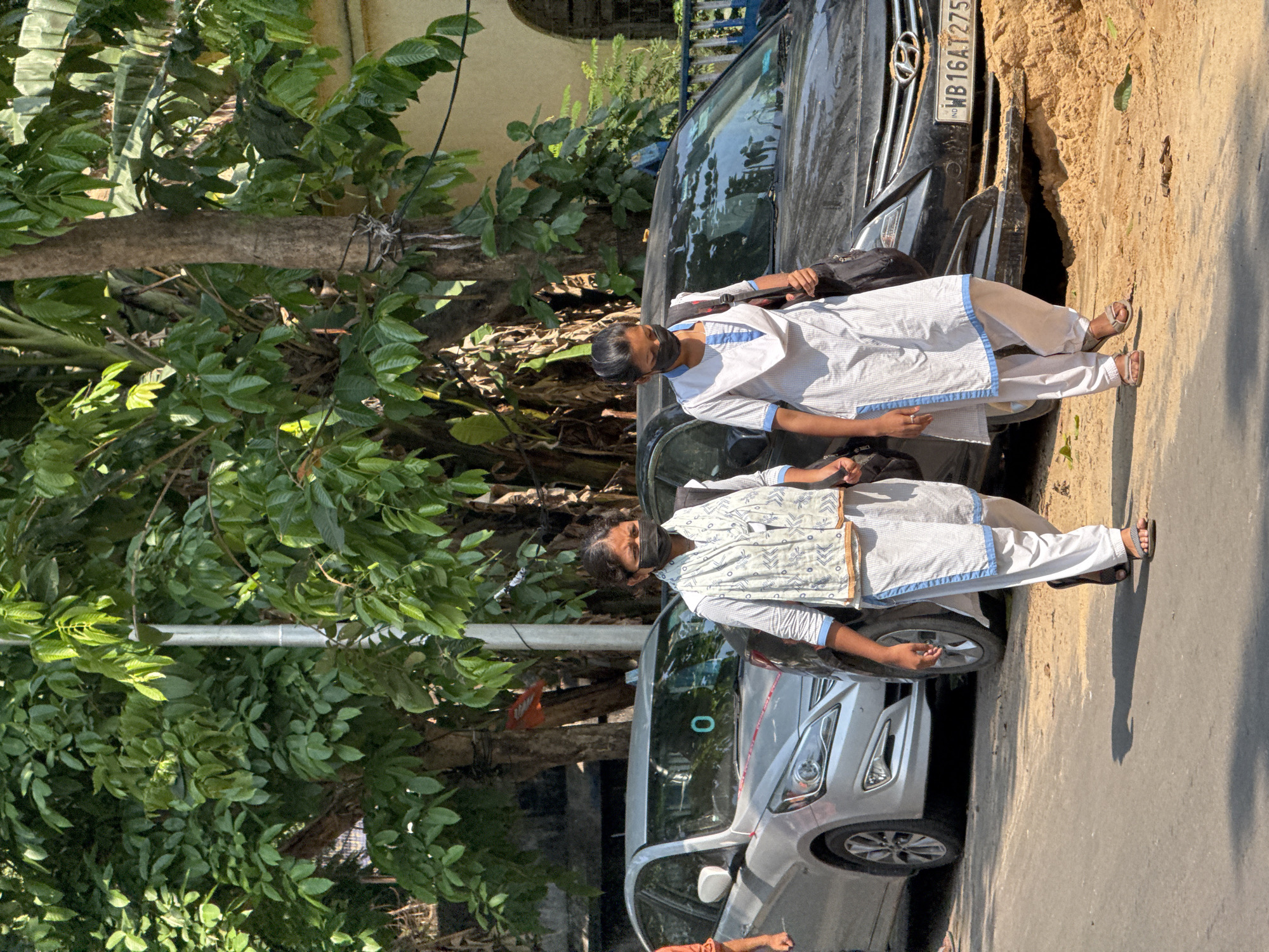 Two females are depicted walking on a paved surface in a street scene in Kolkata, India, during daylight hours. The person on the left wears a light-colored kurta, salwar, and patterned dupatta, with a black face mask. Her right hand is clasped with the left hand of the second individual. The second individual, positioned to the right, wears a white uniform with blue trim, a black face mask, and carries a dark shoulder bag on her left side. She also wears sandals. They are walking towards the viewer's right. Behind them, two parked Hyundai sedans are visible: a silver Verna sedan with a red ribbon on its front and a '0' on its windshield, and a black sedan with West Bengal license plate "WB 16AT2755" partially obscured by a mound of light brown sand/earth. Lush green foliage from multiple trees dominates the background, with a grey utility pole and a portion of a light-colored building with an arched window visible behind the vehicles and trees. The scene is brightly lit, casting shadows on the ground.