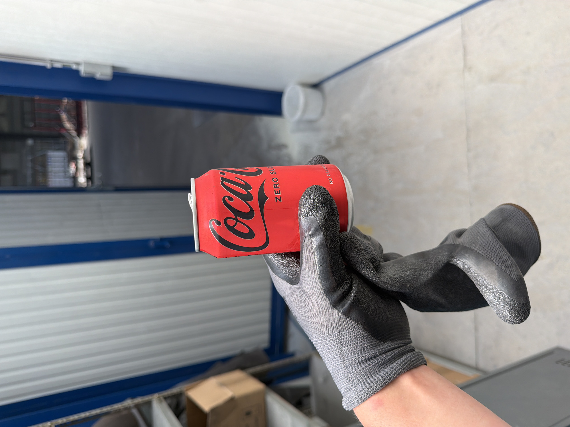 A gloved hand holds a red Coca-Cola Zero Sugar can. The can has the iconic Coca-Cola logo in black script and reads "ZERO SUGAR" below it. The hand is wearing a grey work glove with black textured grip on the fingertips and palm. The background is a light-colored, industrial-looking space with corrugated metal walls and blue trim. In the distance, a large open doorway reveals a bright, blurred outdoor area with what appears to be construction or industrial equipment. A small white bucket is visible on the floor in the middle ground to the right. The overall scene suggests an industrial or storage area, possibly within the city of Mol, Belgium, given the context.