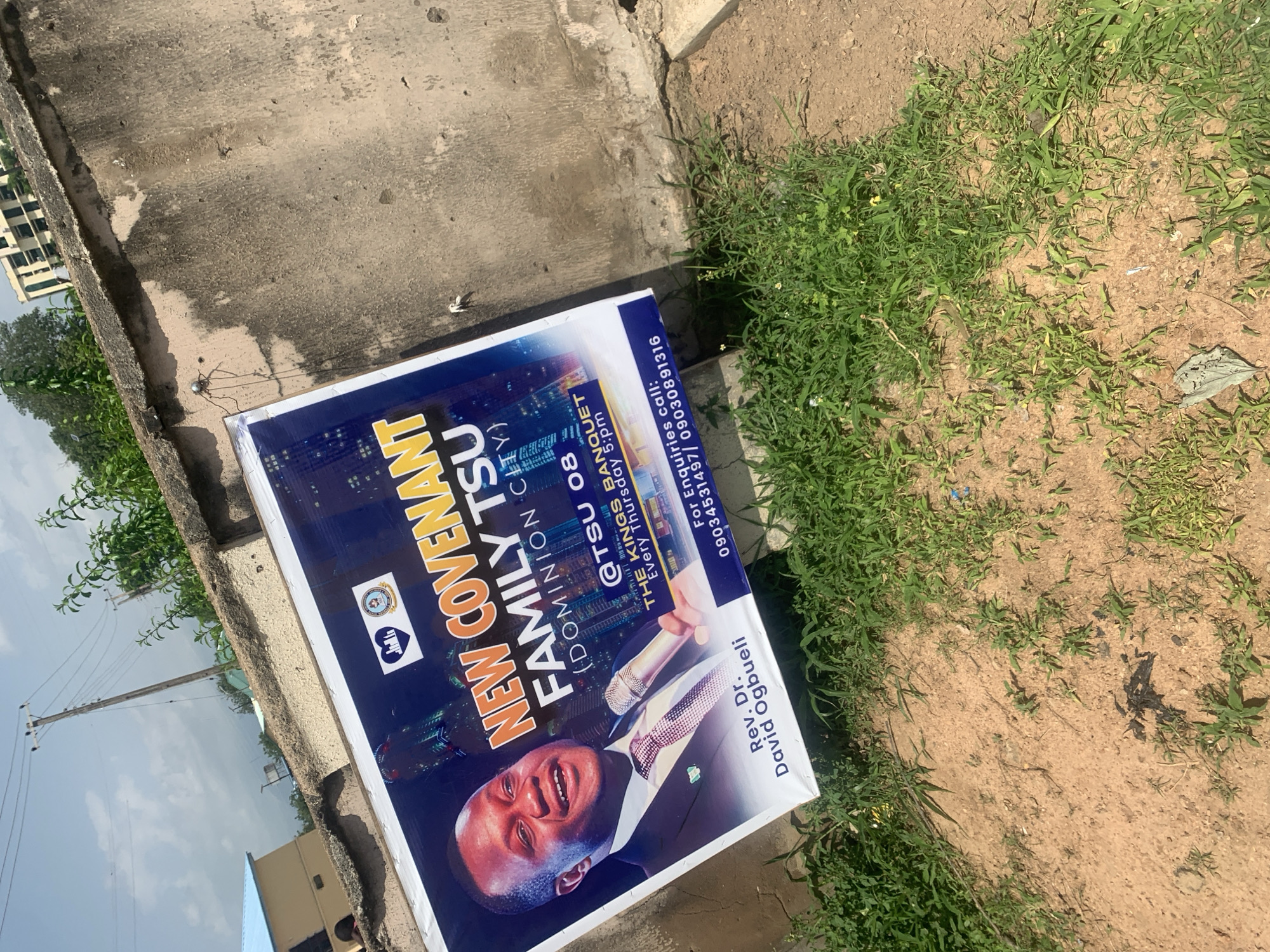 The image shows a poster advertising "NEW COVENANT FAMILY TSU (DOMINION CITY)" with a picture of Rev. Dr. David Ogbueli. The poster is attached to a rough, concrete wall and is partially covered by grass and dirt. It also features the text "@TSU 08 THE KINGS BANQUET Every Thursday 5:00 PM" and "For Enquiries call: 09034531497 / 09030891316". In the background, a light pole with electrical wires and some buildings can be seen under a cloudy sky. The scene appears to be outdoors in Jalingo, Nigeria.