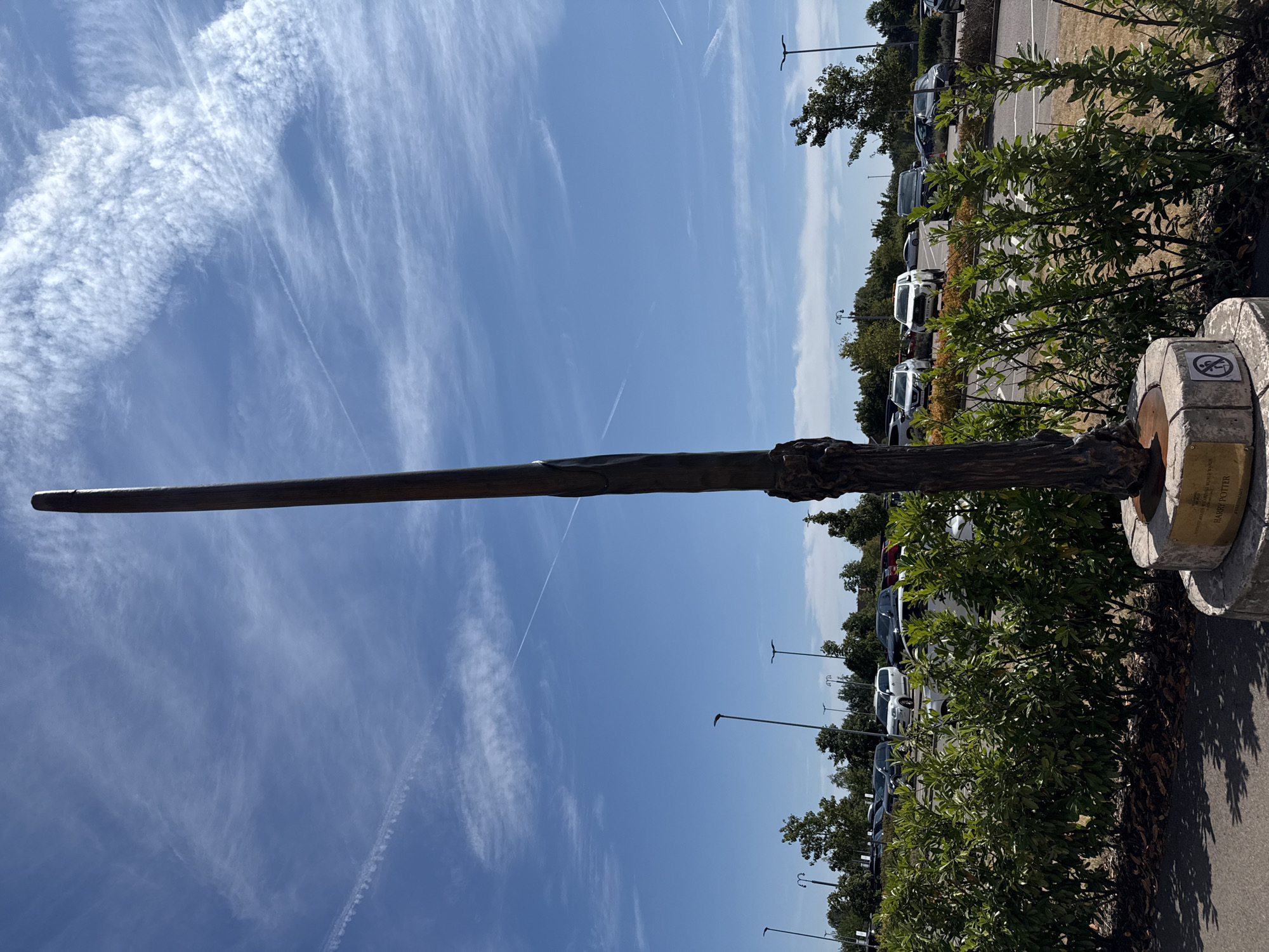 A tall, dark, slender sculpture, resembling a wand, stands vertically on a circular stone base in an outdoor setting. The sculpture has a textured, wood-like appearance and tapers to a point at the top. The base has a plaque with text that appears to read "HARRY POTTER" and "WARNER BROS. STUDIO TOUR LONDON". To the right of the plaque is a circular symbol indicating no climbing.
The sculpture is positioned in front of a low, dense green hedge. Beyond the hedge, a paved area transitions to a parking lot filled with various cars, including white SUVs and darker sedans. In the background, several tall streetlights are visible, along with a line of green trees under a bright blue sky with scattered white clouds and contrails. The lighting suggests it is daytime.
The location is the Warner Bros. Studio Tour London – The Making of Harry Potter, located in Leavesden, near Watford, Hertfordshire, United Kingdom.