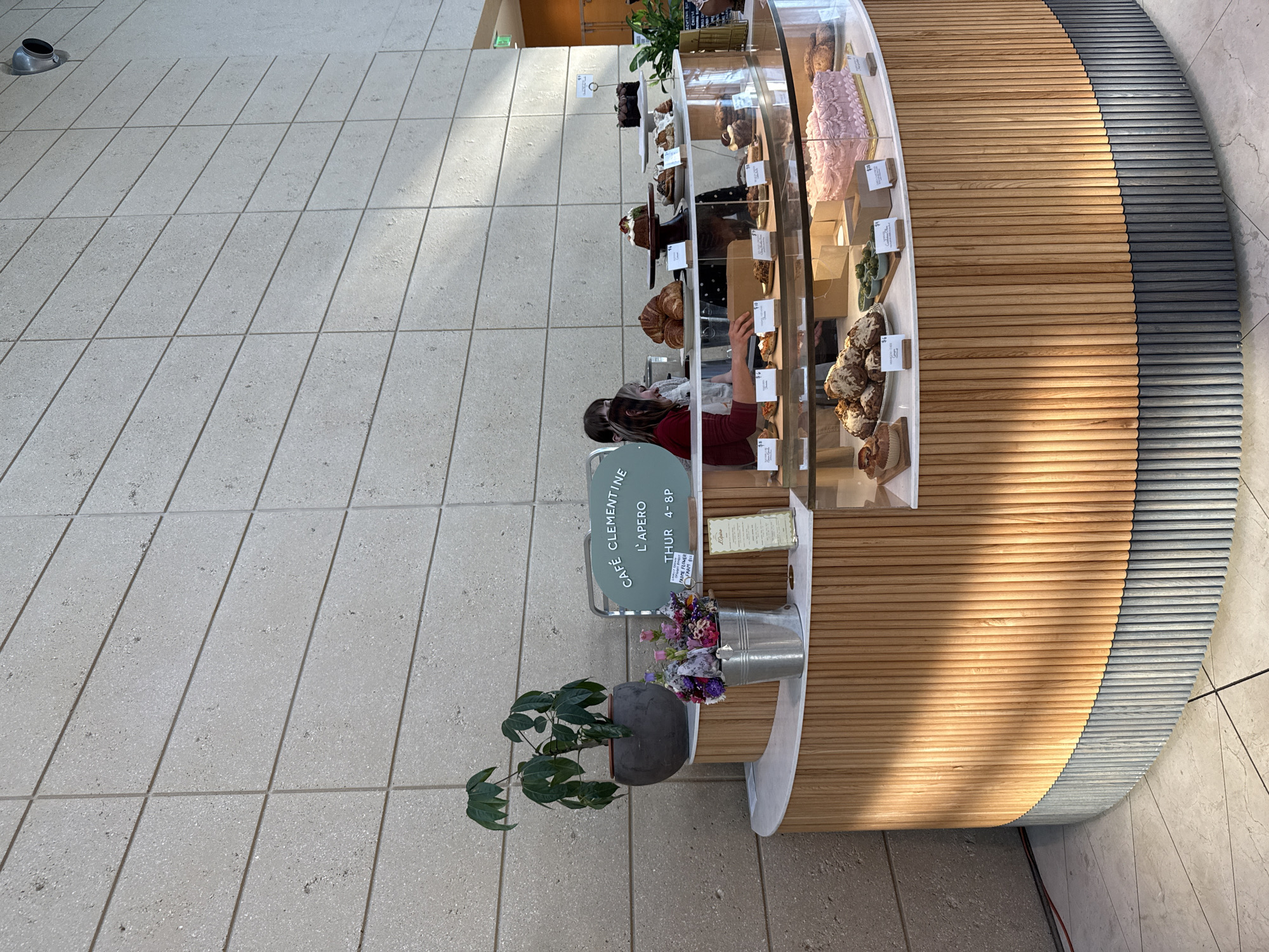 The image displays an indoor scene featuring a modern cafe counter, identified as "CAFÉ CLEMENTINE L'APERO," located within The Dalí Museum in Saint Petersburg, Florida, United States.
The primary subject is a semi-circular service counter with a white marble or stone top. The counter's façade is constructed from vertical wooden slats; the upper section features light-toned wood, while the lower section is composed of darker, possibly grey-painted, wood slats. An integrated curved glass display case in the front of the counter showcases various baked goods, including a pink rectangular cake with decorative piping, several green matcha-like desserts in small bowls, and various scones or similar items, each marked with small white price tags (e.g., "$10", "$8", "$6").
On the upper surface of the counter, additional pastries are displayed, including croissants and multiple cakes on serving stands (one bundt cake and one dark chocolate cake). To the left, a metallic bucket holds a colorful bouquet of flowers. Behind this, a potted plant with large green leaves rests on a raised, white-topped platform. A light green oval sign prominently displays "CAFÉ CLEMENTINE L'APERO THUR 4-8P". A smaller menu board labeled "L'Apéro" is also visible.
Two individuals are positioned behind the counter. The most visible person wears a reddish-brown long-sleeved top and a light-colored apron, with light brown hair. This individual is seen from the chest up, smiling slightly while appearing to attend to items on the counter. A second person is largely obscured behind the first, wearing a dark top and apron.
The background consists of a tall wall composed of large, light-grey rectangular stone or concrete blocks with visible horizontal grout lines. To the far right, a narrow orange wall segment and an illuminated green "EXIT" sign are present, indicating a doorway or passage. A cylindrical silver fixture is mounted on the upper right wall. The floor is covered with light-colored, possibly beige, square tiles laid diagonally. Natural light illuminates the scene from the right, casting distinct shadows on the counter's wooden slats.
