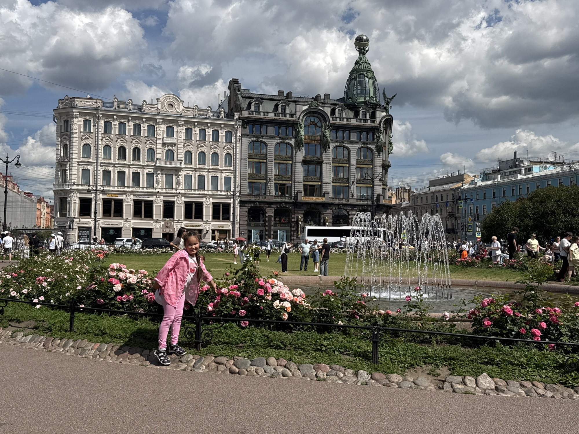 The image depicts a wide shot of a public square in Saint Petersburg, Russia, likely Nevsky Prospect, given the architectural style of the buildings. In the foreground, a young girl in pink pants and a pink jacket stands by a vibrant rose bush, leaning on a black metal fence. She has a backpack and is looking towards the camera with a smile. Behind her, a large, ornate fountain sprays water into the air.
The background features several grand, historic buildings. The most prominent is the Singer House, characterized by its distinctive green dome topped with a globe. Adjacent to it is a lighter-colored, classical-style building with many windows. Further in the distance, more buildings and a white bus are visible. The sky is filled with dramatic, fluffy clouds. Several other people are scattered throughout the square, some walking, others standing near the fountain. The overall scene suggests a sunny, pleasant day in a bustling city center.