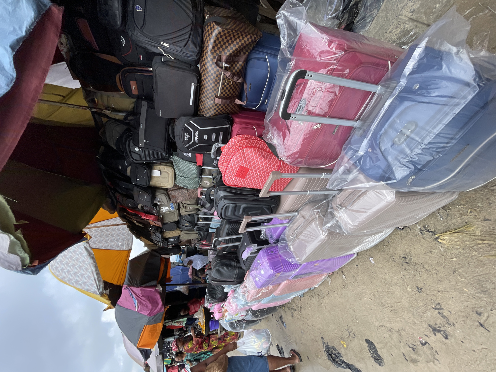 The image captures a bustling outdoor market scene in Lekki, Nigeria, focused on a stall displaying a wide array of luggage. In the foreground, a row of suitcases in various sizes and colors, including purple, pink, brown, and red, are neatly arranged, many of them covered in protective plastic. Behind the suitcases, a collection of backpacks and duffel bags are stacked on shelves and hung on display, featuring brands like "HPILLO" and "LACOSTE." Several people are visible in the background. A woman wearing a colorful patterned dress and a headscarf is interacting with a vendor. Another individual, dressed in a white head covering, appears to be a customer. The market stall is covered by colorful umbrellas and tarps, suggesting an open-air setting. The ground is a mix of dirt and patchy pavement, with some debris visible. The overall atmosphere is one of lively commerce and a diverse selection of travel goods.