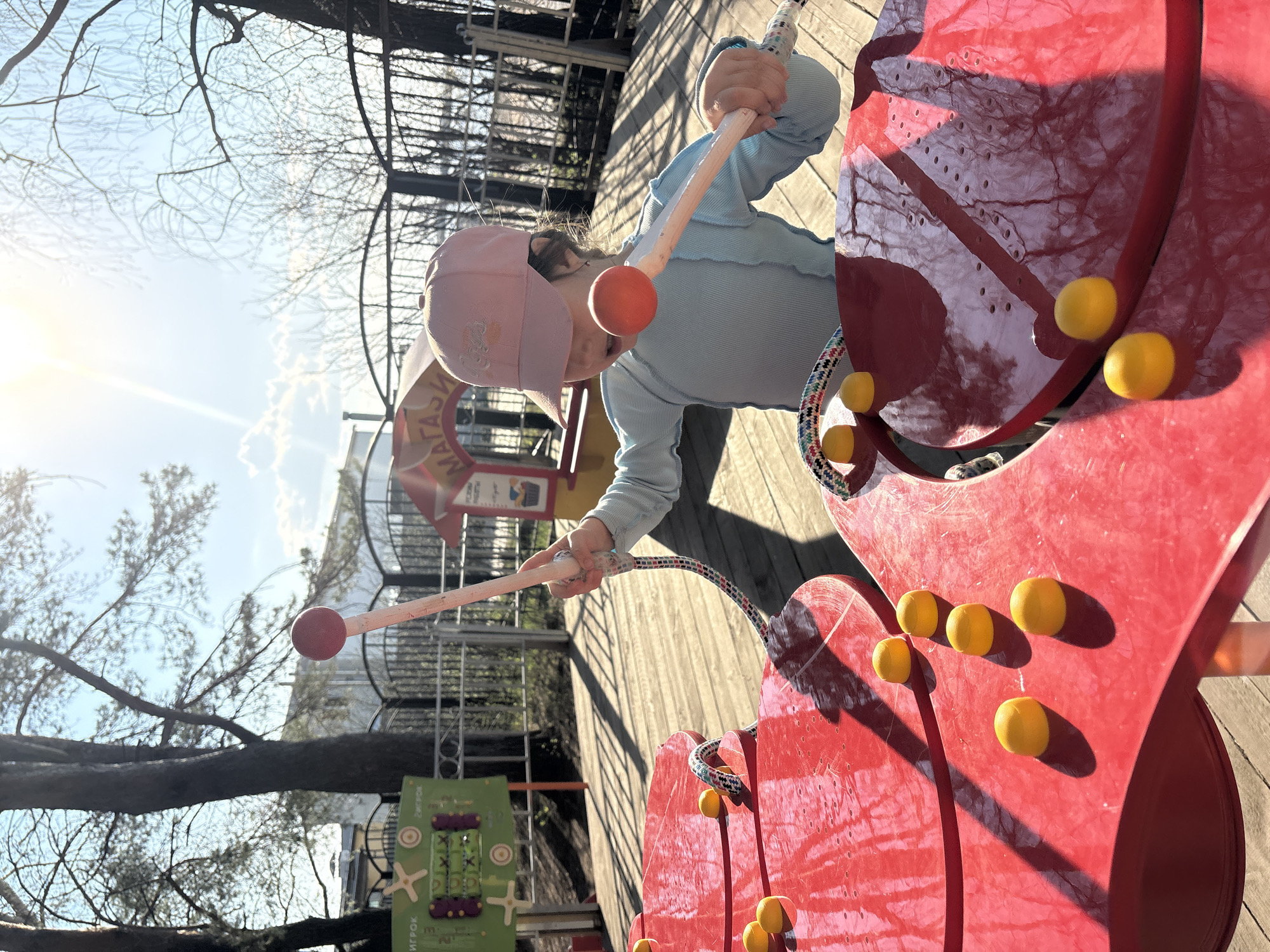 A child in a light blue long-sleeved shirt and pink hat plays on a red musical instrument in what appears to be a playground. The instrument has yellow ball-shaped protrusions. The child holds two mallets, each with a red ball at the end. The background includes trees with bare branches, a metal fence, and other playground equipment. The location is in the city of Chaikovsky, Russia.