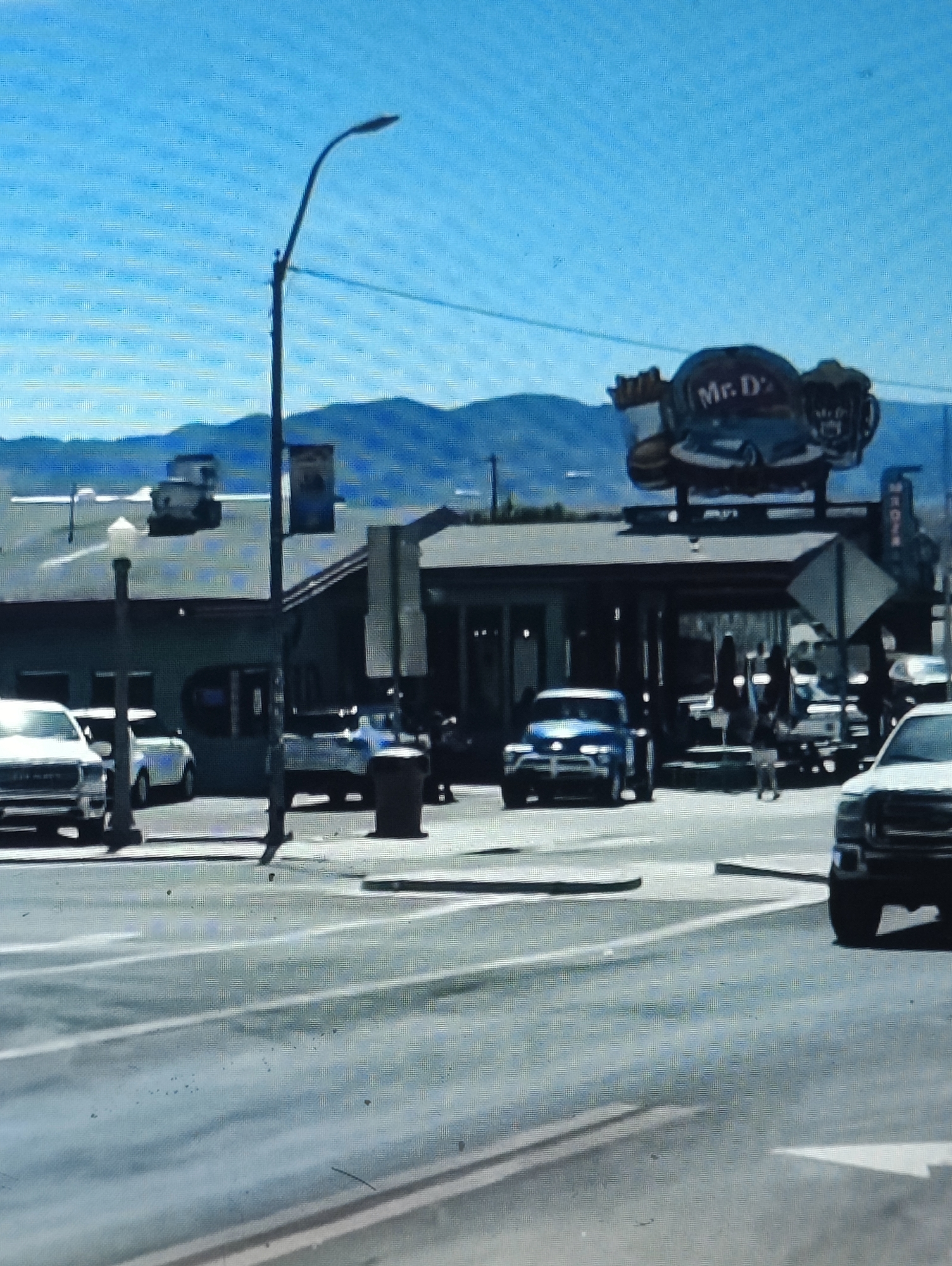 The image captures a street scene with a building in the background that appears to be a restaurant or drive-through establishment with a prominent sign that reads "Mr. D's". Several vehicles are present, including a white pickup truck in the foreground to the right, a blue sedan in the middle ground, and other vehicles parked or driving on the street. A street lamp is visible on the left side of the frame. The environment is outdoors with a clear blue sky and mountains in the distance, suggesting a dry, sunny day. The image quality is somewhat grainy, with a noticeable digital artifact pattern.