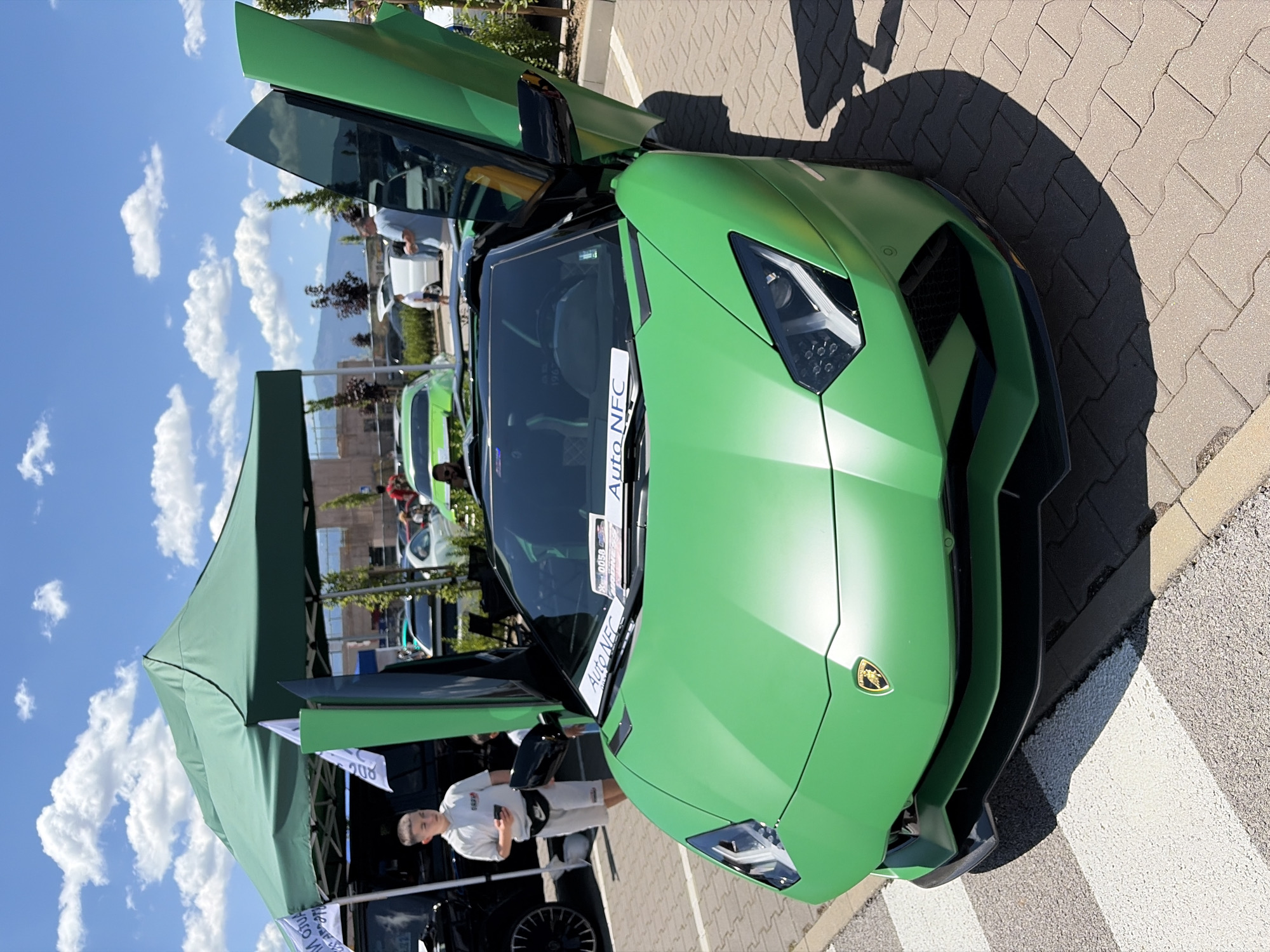 A green Lamborghini Aventador is parked on a paved lot, possibly at an outdoor event or car show. The car's dihedral doors are open, revealing the black interior. A white banner with "Auto NFC" printed on it is attached to the front of the car. In the background, several other cars are visible, along with a green tent and people milling about. The sky is blue with scattered clouds. The setting appears to be an urban environment on a sunny day.