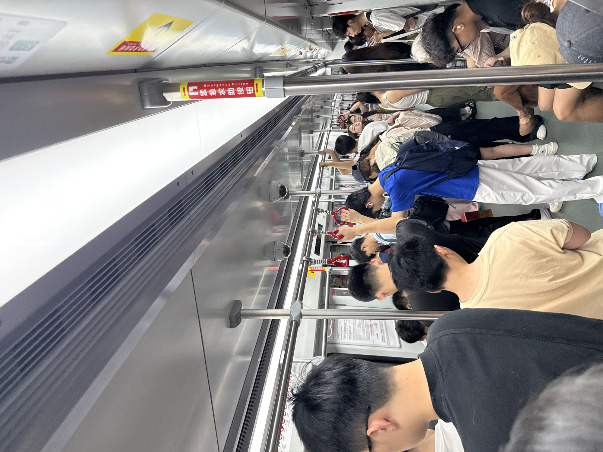 The image depicts the interior of a crowded subway car, likely in Haizhu District, China. Passengers are standing and sitting, holding onto poles and overhead bars. The car features metallic walls and ceiling, with fluorescent lighting. Several safety signs and emergency buttons are visible, including a red and yellow sign with Chinese characters and the word "Emergency button" in English. People of various ages and genders are present, dressed in casual attire. Some individuals are looking at their phones, while others appear to be conversing or simply enduring the crowded conditions. A security camera is mounted on the ceiling. The overall atmosphere suggests a busy commute during peak hours.