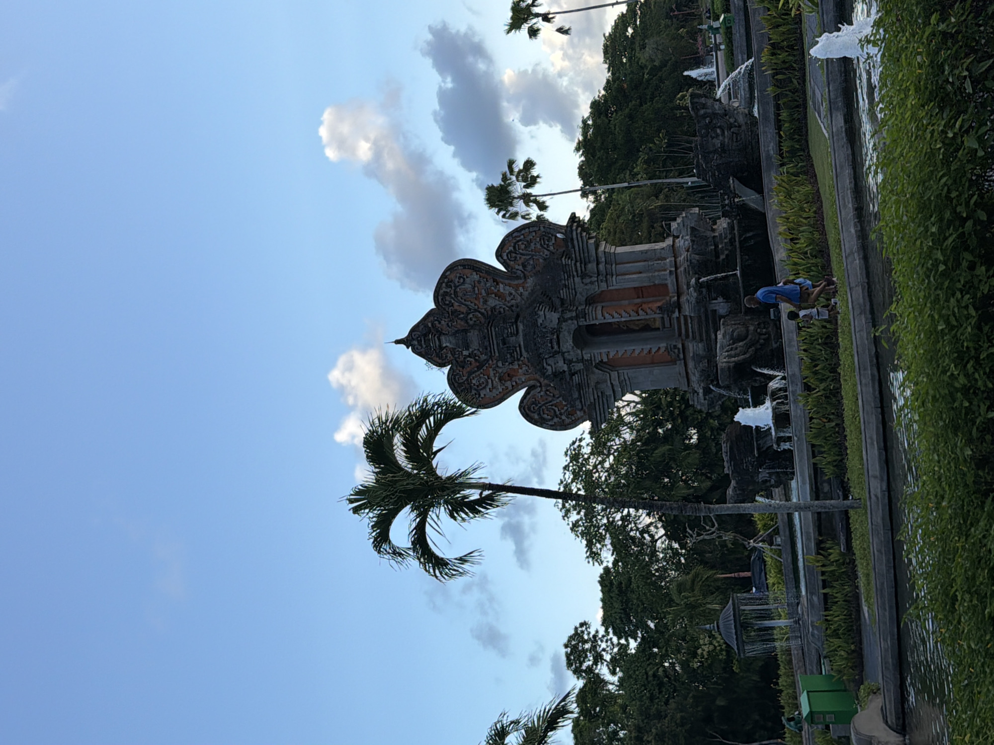 This is an image of the Bajra Sandhi Monument in Kuta Selatan, Bali, Indonesia. The monument is a large, ornate structure with intricate carvings, resembling a temple. It is surrounded by lush greenery and water features, including fountains. In the foreground, two people, a man and a child, are walking on a pathway. The sky is blue with some scattered clouds. The image captures a serene outdoor scene, likely in the late afternoon or early evening, given the lighting.