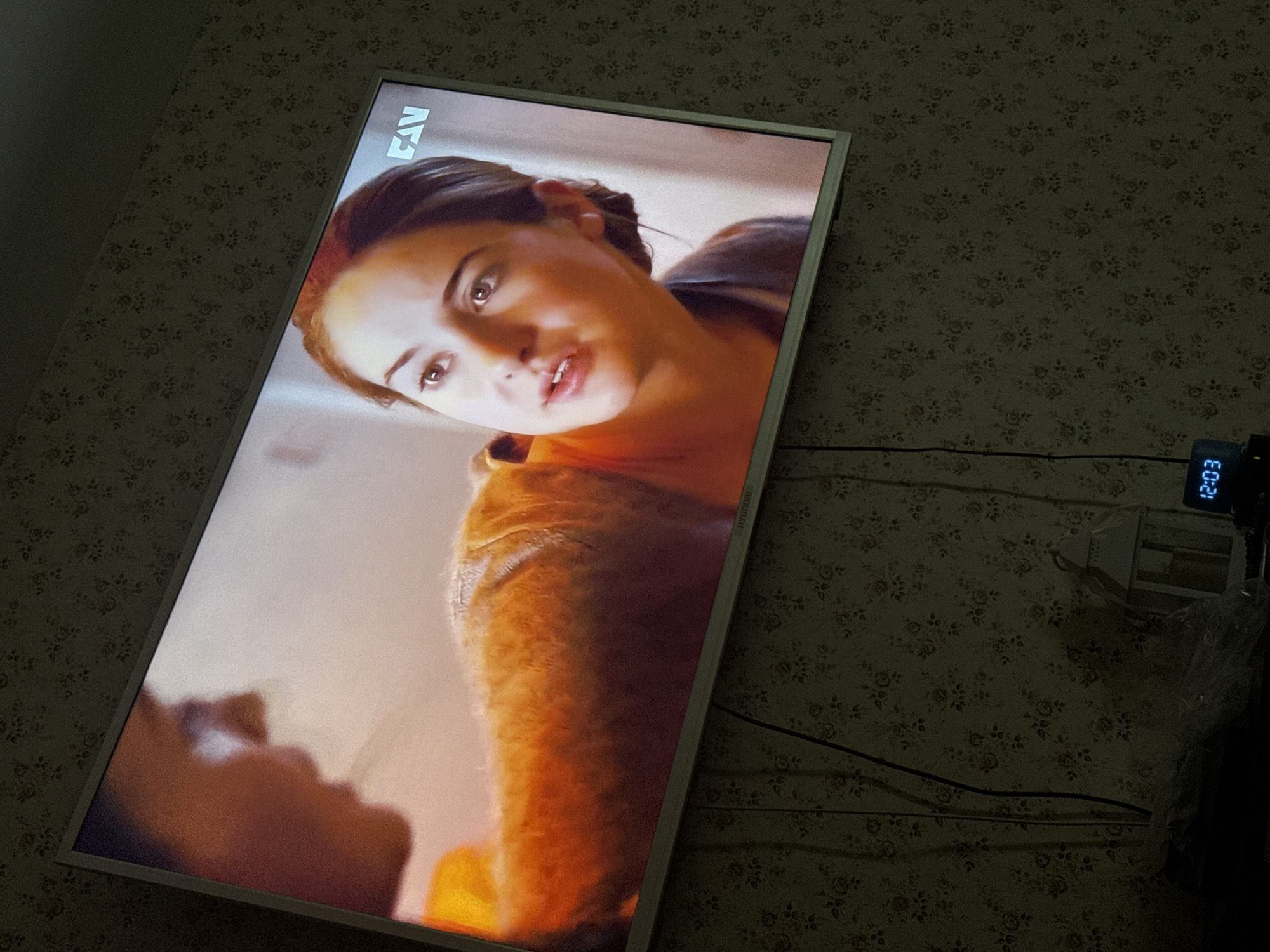 The image shows a television screen displaying a woman's face in close-up. The television is mounted on a wall with floral wallpaper. To the right of the television is a digital clock displaying the time "12:03." The wallpaper pattern consists of small, repeating floral motifs. Cords and an electrical outlet are also visible on the wall to the right of the television. The location is in Mytishchi, Russia.