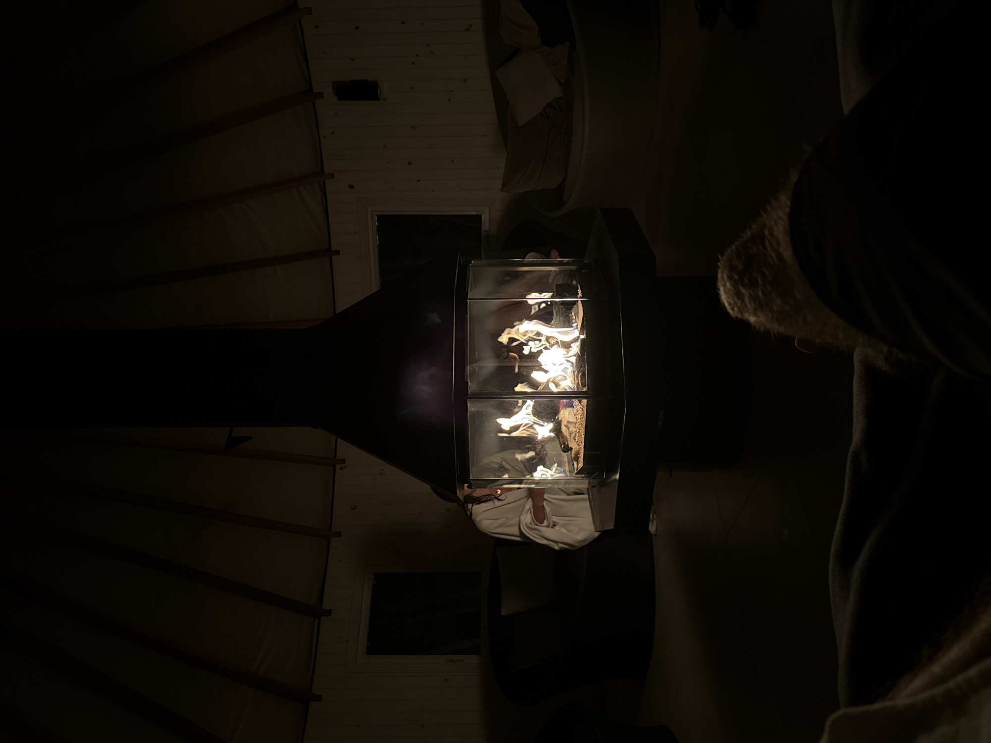 A low-angle interior shot depicts a modern, glass-enclosed fireplace. The fireplace is central, and its flames illuminate the otherwise dimly lit room. To the upper-left, structural elements of a rounded yurt ceiling are visible, while a couch sits to the upper-right. A person with a fur-lined hood is partially visible in the lower-right. The location is not explicitly visible and therefore cannot be confirmed in Montreal, Canada.