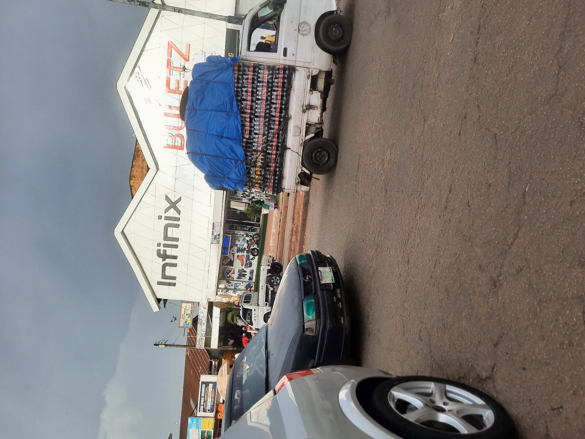 The image shows a street scene in Jos, Nigeria. The sky is overcast. A white truck heavily laden with bottled drinks covered with a blue tarp is parked on the side of the road. The truck is positioned in front of a white building with "Infinix" and "BULETZ" signs. Two cars are parked in front of the truck, a dark-colored one and a silver one. A few people can be seen in front of the Infinix building. The street is paved. Utility poles and additional signage are visible in the background.