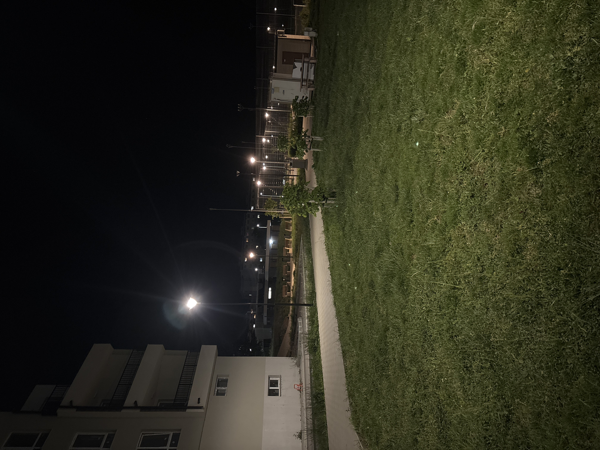The image depicts a nighttime scene, seemingly in a residential area. On the left is a light-colored building, possibly an apartment complex, with visible windows and external staircases. A street lamp illuminates the scene. Adjacent to the building is a paved walkway. To the right of the walkway is a large expanse of grass. In the background, there is a row of streetlights and indistinct structures, possibly related to a park or communal area. The scene lacks human presence. There is no definitive evidence to confirm the location within Burgas, Bulgaria.