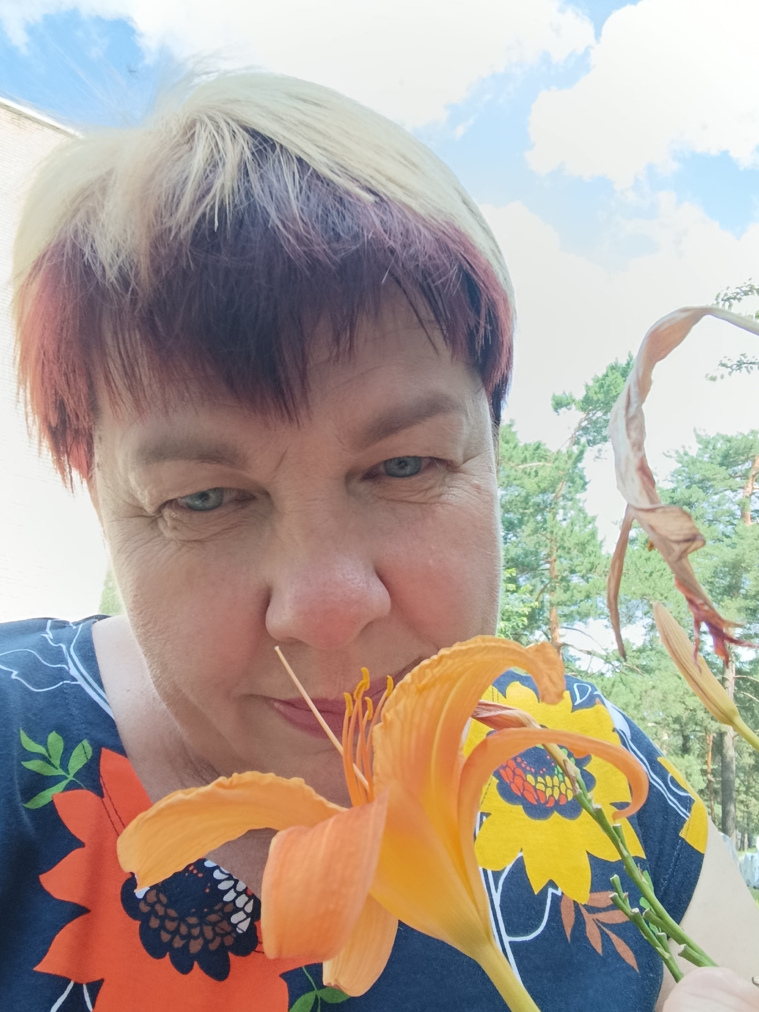 A woman with short, blonde and reddish-brown dyed hair is taking a selfie outdoors. She is wearing a dark blue shirt with a colorful floral pattern. An orange lily is held up close to her face, partially obscuring her mouth and nose. The background shows a blue sky with white clouds and green trees. The scene appears to be in a park or garden setting in the city of Svietlahorsk, Belarus.