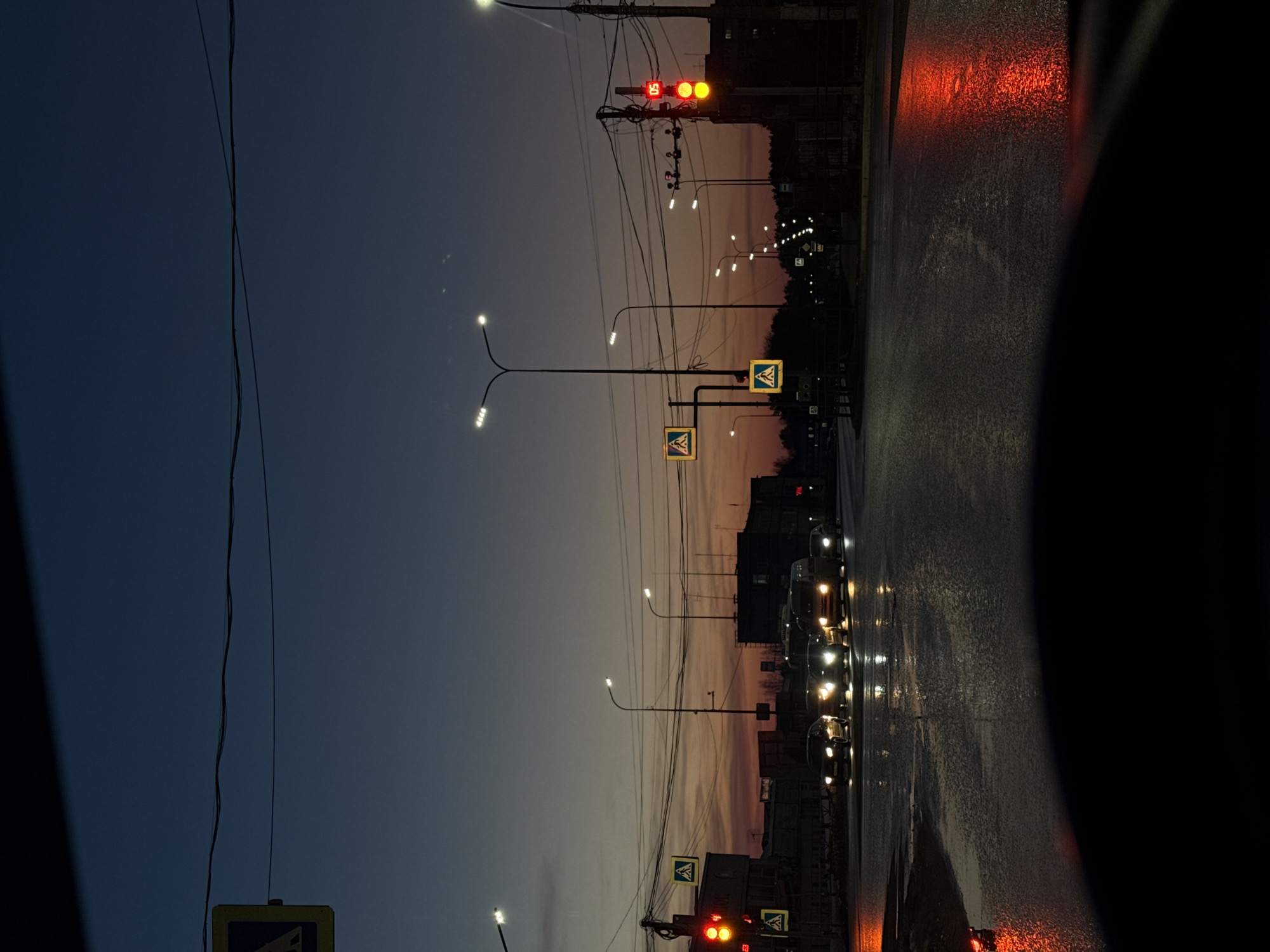 The scene depicts a wet urban intersection in Chaikovsky, Russia, during dusk or night, viewed from the interior of a stationary vehicle. The asphalt road surface is dark, wet, and highly reflective, mirroring the illuminated light sources. Ahead, approximately five to six vehicles are stopped with their headlights active, awaiting a signal.
Two prominent traffic light poles are visible. The pole on the left displays two active red circular lights. The pole on the right also displays two active red circular lights, with a digital countdown timer above them showing "05." Multiple yellow square pedestrian crossing signs, featuring a black walking figure, are affixed to various poles across the intersection.
The environment is illuminated by numerous active streetlights on tall, black poles, casting white light. An extensive network of overhead utility wires crisscrosses the sky, connected to several utility poles. Dark silhouettes of multi-story buildings are visible in the background, partially obscured by the urban infrastructure. The sky transitions from a deep dark blue at the zenith to a gradient of orange and purple hues near the horizon, indicative of twilight. The lower edge of a vehicle's dashboard and windshield is visible in the foreground, with red reflections appearing on the left side of the windshield. No individuals are discernible within the frame.