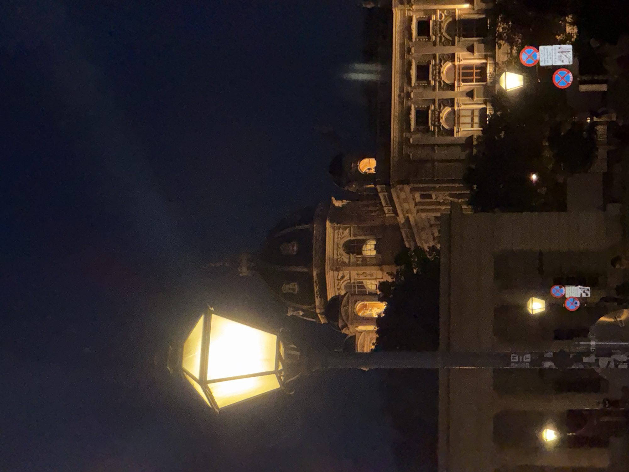 The image depicts a nighttime view of the Natural History Museum (Naturhistorisches Museum) in Vienna, Austria. A prominent street lamp with a glowing yellow light stands in the foreground, its ornate pole covered in stickers. Behind the lamp, the museum's grand facade is illuminated, showcasing its architectural details, including arched windows and decorative elements. A large dome is visible on the museum's roof. In the mid-ground, a lower building with arches can be seen. Several smaller street lamps and traffic signs, including "no parking" signs, are visible in the lower portion of the image. Trees are scattered throughout the scene, and a few indistinct figures can be observed in the background, suggesting a public space at night. The sky is dark and clear.