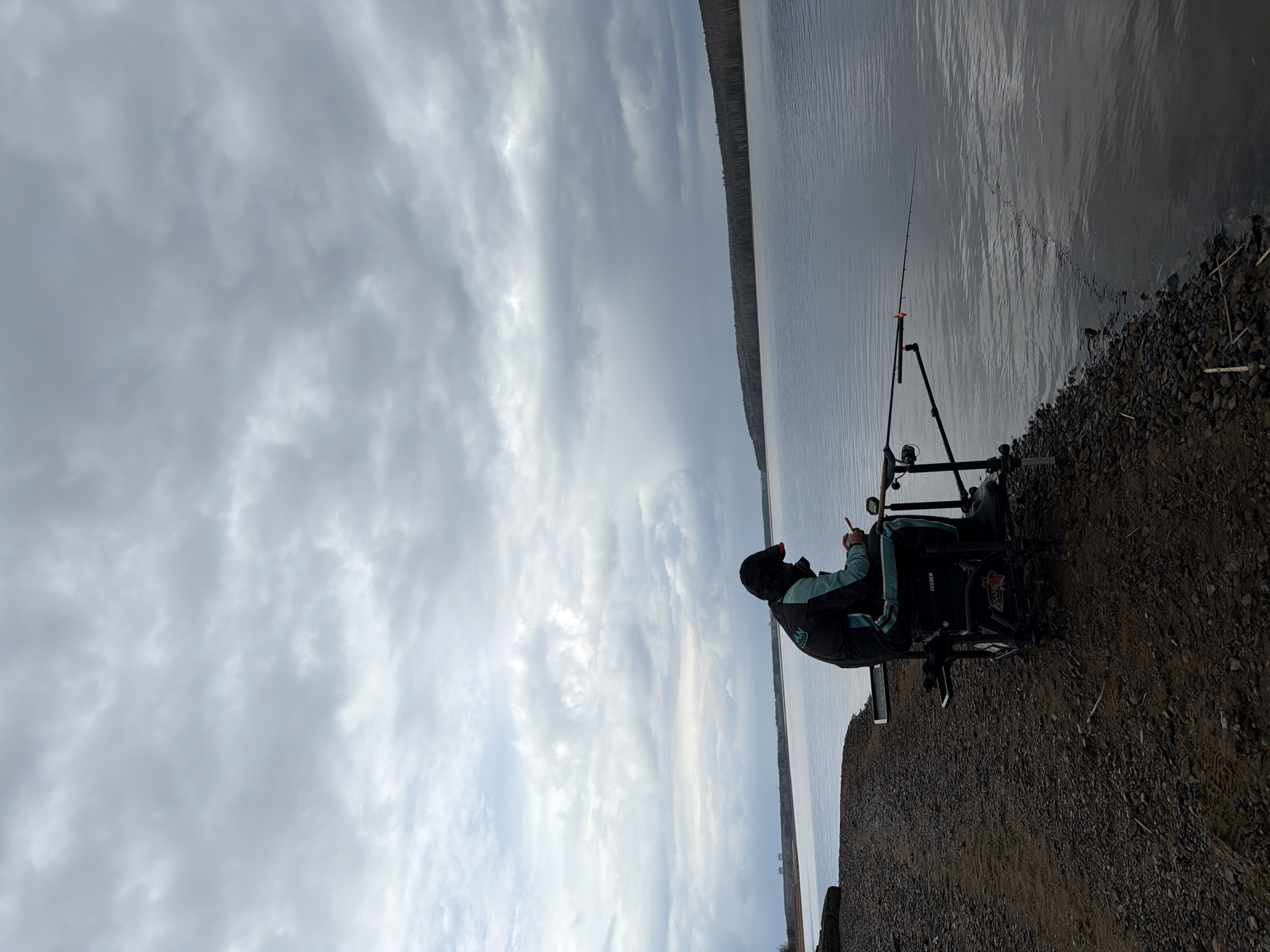 A man is fishing on the banks of the Kama River in Chaikovsky, Russia. He is sitting in a fishing chair with a fishing rod placed on a rod rest. The river stretches out to the horizon under a cloudy sky. The river bank is rocky.