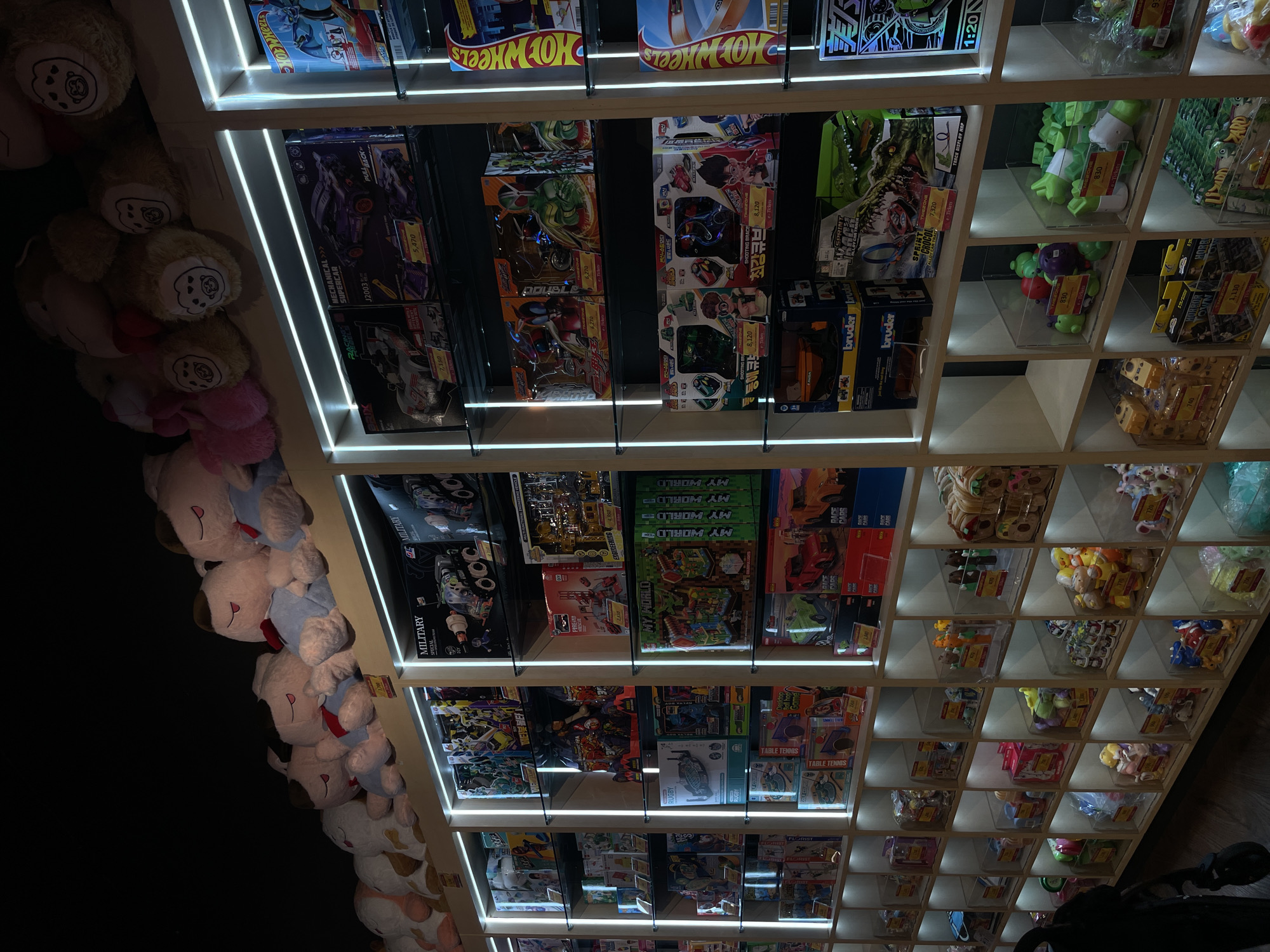 The image depicts a retail store, possibly a toy store, in Manado, Indonesia. The store is filled with shelves displaying a variety of toys and plush items. On the top shelves, numerous large, plush stuffed animals are arranged, some with distinct facial features and wearing clothing. Below these, various toy boxes are neatly organized on illuminated shelves. These boxes contain items such as model cars, construction sets, and action figures. The lighting in the store is predominantly from white LED strips that illuminate the shelves, creating a bright and organized display. The shelves themselves are made of a light-colored material, likely wood or laminate. Towards the bottom of the image, smaller bins containing loose toys, such as small plastic figures and balls, are visible. The overall impression is that of a well-stocked toy store with a modern display setup.