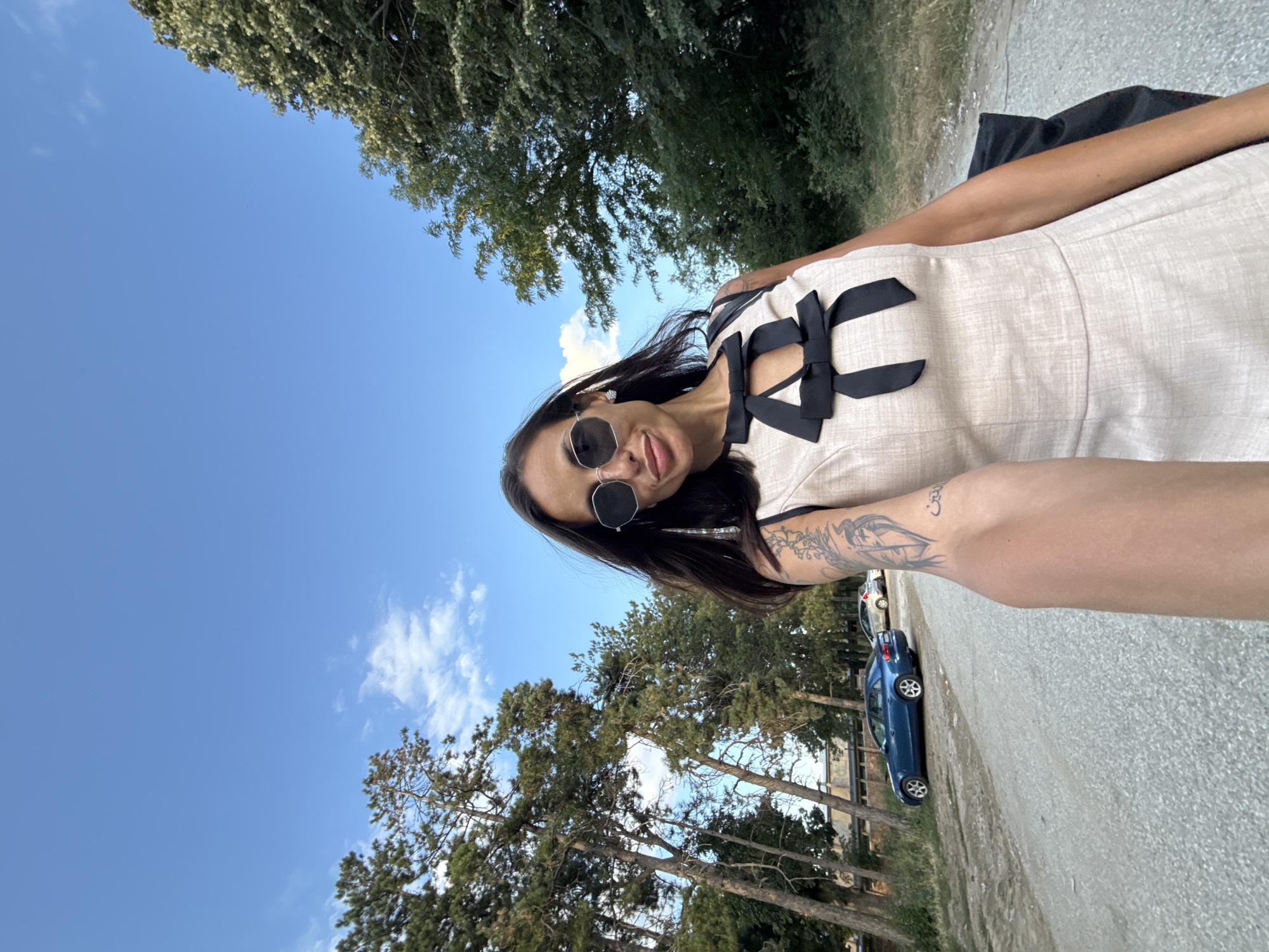 A woman is taking a selfie outdoors. She is wearing sunglasses, a light-colored dress with black bows, and has tattoos on her arm. Behind her, a dirt road is visible with a blue car parked on the side, and trees line the road. The sky is blue with some white clouds. The location is the city of Pleven, Bulgaria.