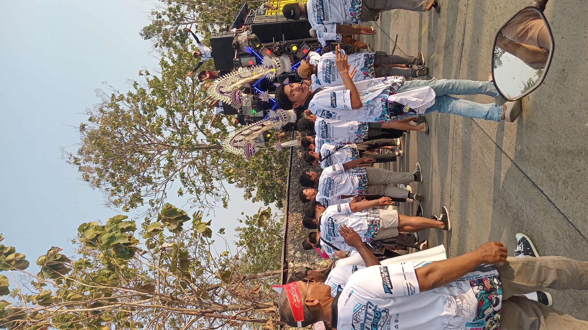 This image captures a scene in Tinapan, Indonesia, where a group of young men in matching white t-shirts are gathered on a paved road. The t-shirts feature colorful graphics and text, suggesting they are part of a team or organization. Some individuals are walking, while others are standing and observing. In the background, a large speaker system is set up, adorned with decorative elements that appear to be part of a procession or performance. Trees with green foliage line the background, and a stone wall is visible behind the crowd. The sky is clear and bright, indicating it is daytime. The overall impression is that of a community event or celebration.