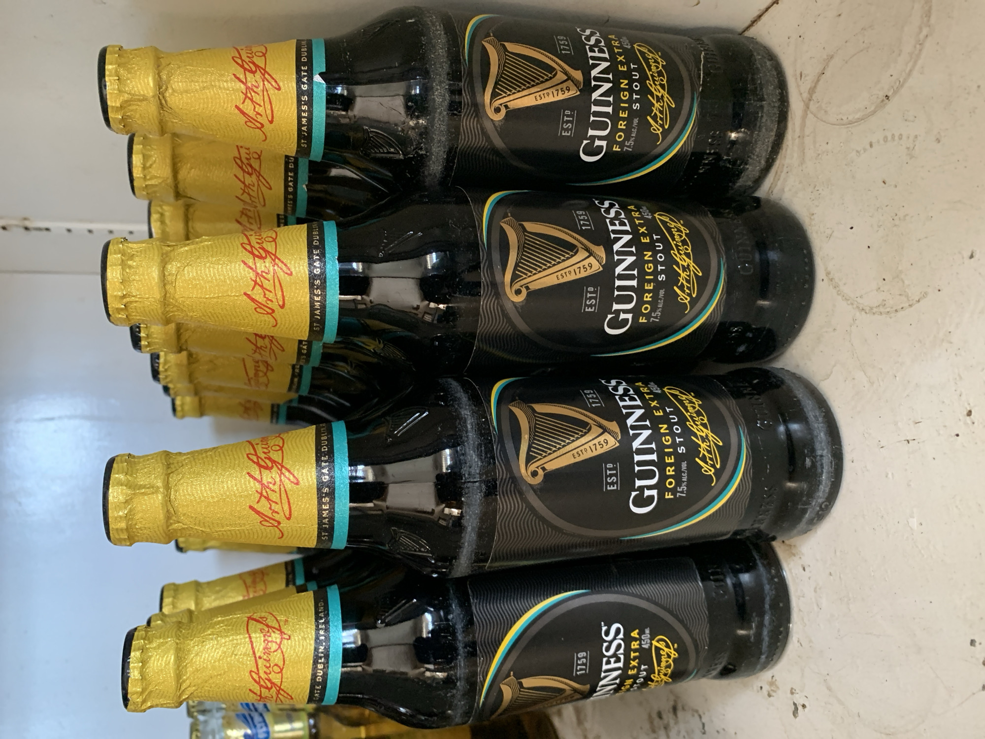 The image shows a shelf or storage area containing multiple bottles of Guinness Foreign Extra Stout. The bottles are dark and have gold foil coverings around the neck with red lettering and a turquoise band below. The labels on the bottles prominently display the Guinness logo and the words "FOREIGN EXTRA STOUT" with the alcohol content listed as 7.5% alc/vol. They appear to be in a fridge or cold storage. The bottles are dusty or have some residue on them. The city of New Stadium, Nigeria is not detectable in the image.