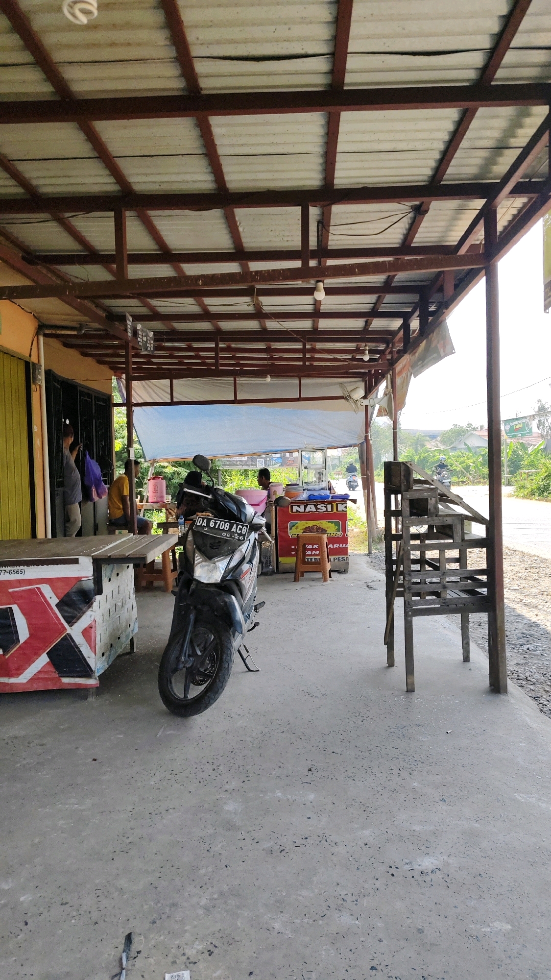 The image depicts an outdoor scene at what appears to be a roadside food stall in Banjarmasin, Indonesia. A metal-framed roof supported by wooden poles covers a paved area. In the foreground, a dark-colored motorcycle is parked on the concrete. To the left, a table with a red, white, and black patterned facade is visible, with a person partially obscured behind it. Further back, a food cart is set up with a sign that reads "NASI K." Several people are seen near the food cart, one of whom is sitting. A wooden structure, possibly a bench or shelving unit, stands to the right. The background shows a street with passing motorcycles and lush green vegetation, suggesting a semi-urban or peri-urban environment. The overall impression is that of a casual, open-air eating establishment in a tropical setting.