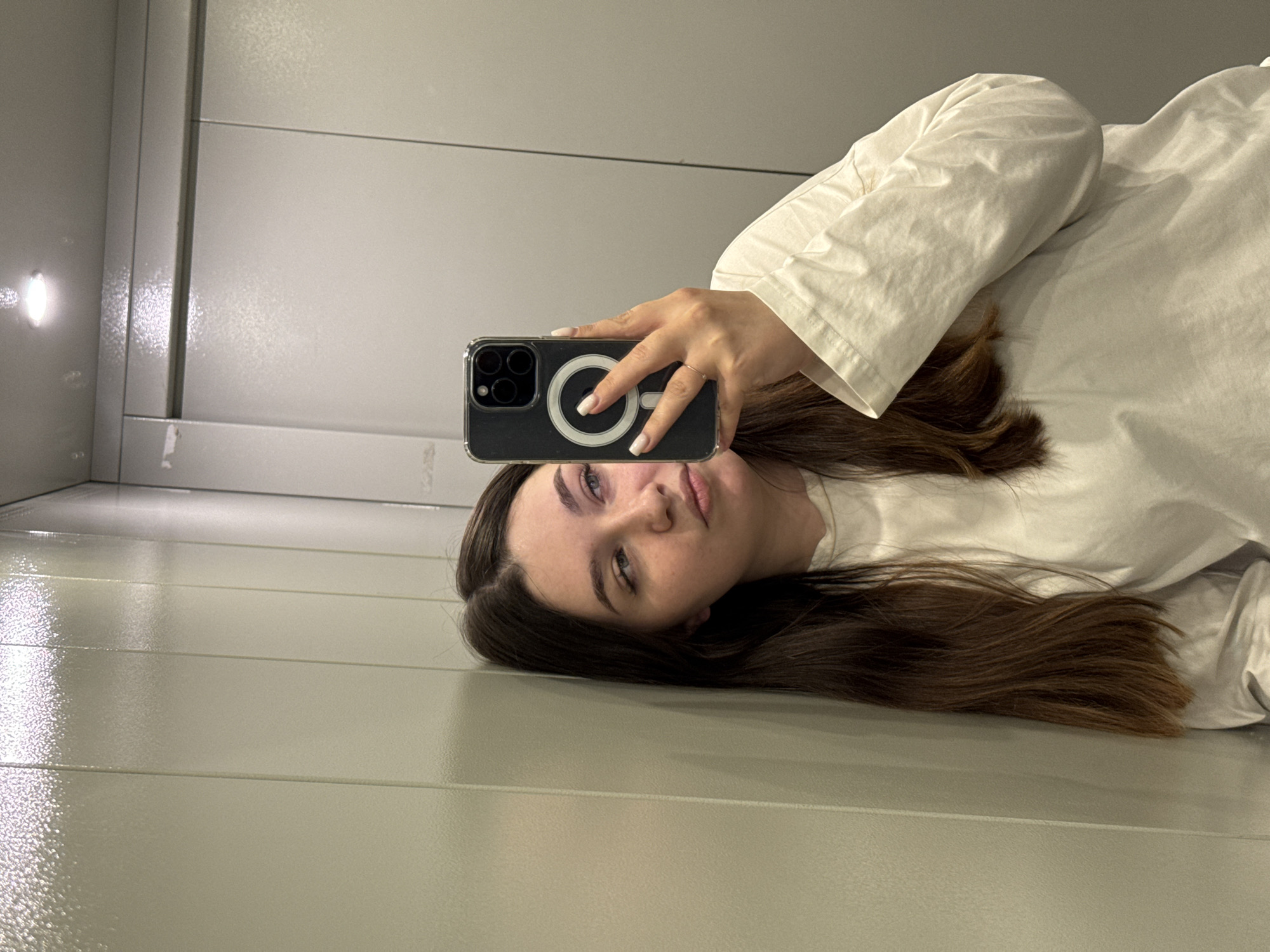 A young woman with long brown hair and wearing a white shirt is taking a selfie in an elevator. She holds a black iPhone in her right hand with a ring on her finger. The elevator has gray walls with horizontal ridges. A light fixture is visible in the upper left corner of the elevator. The location is Dikaya Gar', Russia, based on provided information, though this detail is not directly discernible from the image itself.