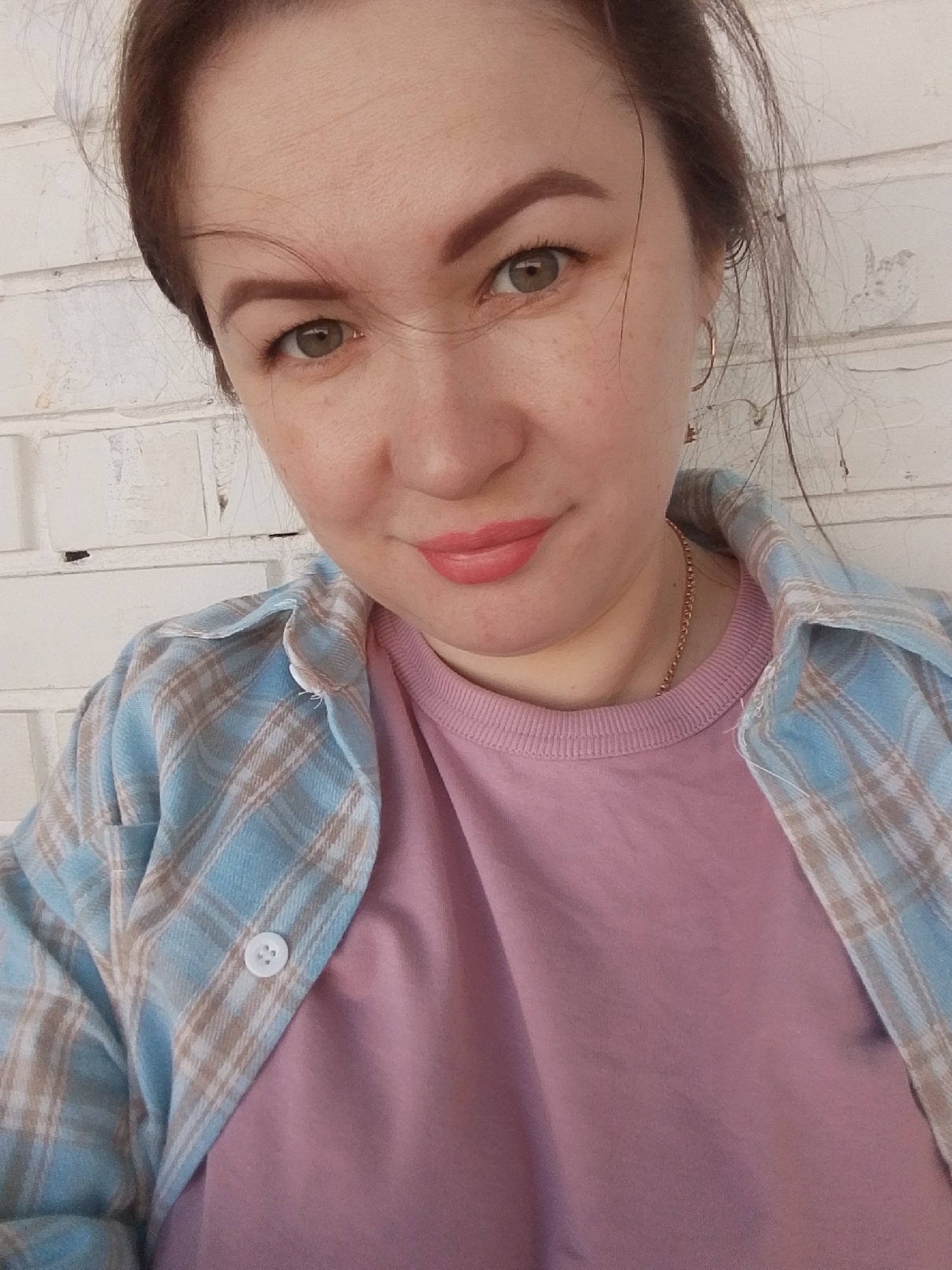 A woman is pictured in a close-up shot. She has fair skin, brown hair pulled back, and green eyes. She's wearing a light pink t-shirt underneath an unbuttoned blue and tan plaid flannel shirt. A thin gold necklace is visible around her neck. The background features a white brick wall. There's no information to confirm the city of Noviy, Russia as her location.