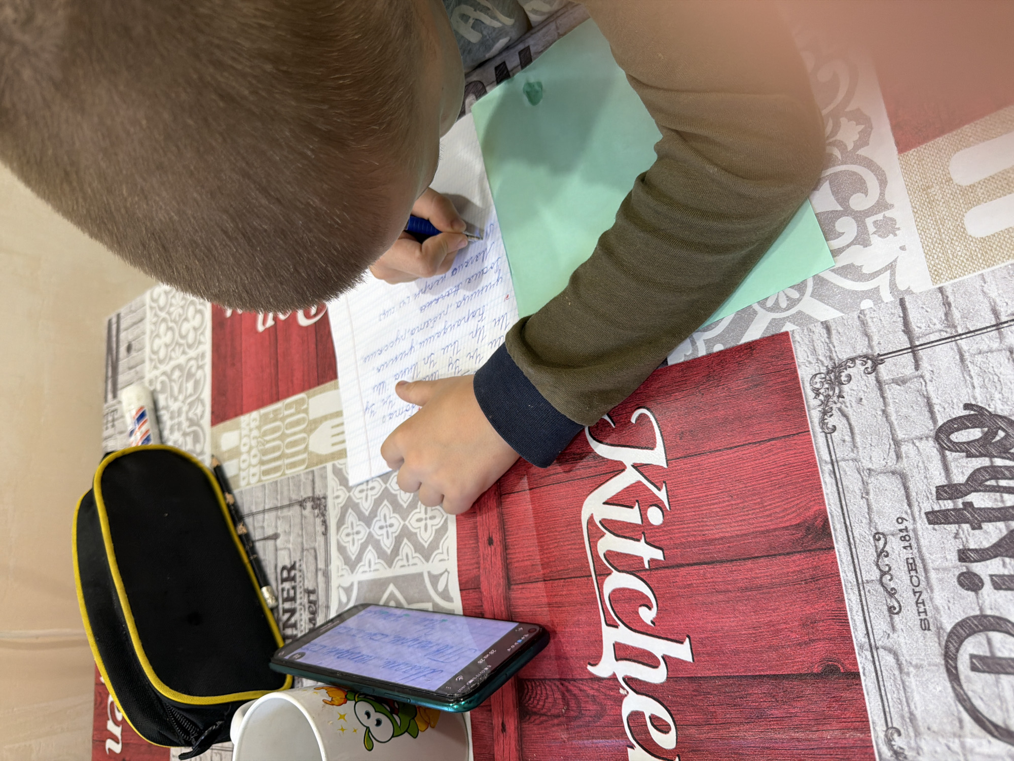 A child is writing on a piece of lined paper on a table. He is using a blue pen. A green sheet of paper is next to the paper he is writing on. A pencil case, a pencil, a phone, and a mug are on the table. The table has a patterned surface. The image contains the text "SINCE 1819 Kitchen". The child is writing in Russian. The text on the page includes "Kapurdawu yserma yzema, ученица, ребята, русские". The objects are located on a table with a decorative "Kitchen" motif in Agoy, Russia.