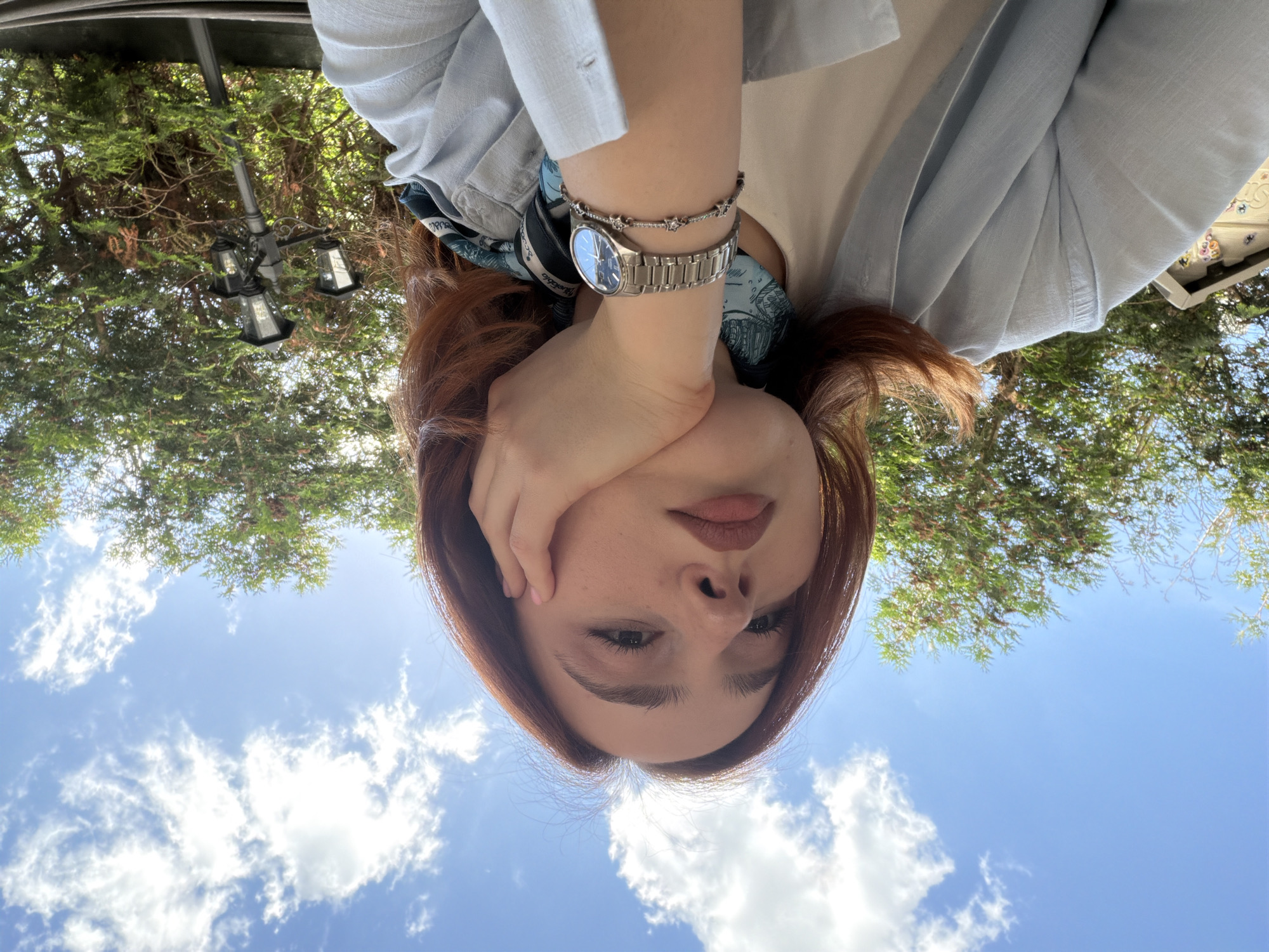 An outdoor scene in the city of Strasbourg, France, features a female subject depicted from a low angle, gazing directly towards the camera. The subject has auburn hair, a fair complexion exhibiting freckles, and wears a light blue collared shirt over a light-colored top. A patterned blue, white, and black scarf is visible at her neck. Her right hand, displaying light pink nail polish, supports her chin. On her left wrist are a silver-toned metal watch with a blue dial and a separate silver-toned bracelet featuring star-shaped charms. The background consists of a bright blue sky with scattered white cumulus clouds, and dense green foliage from trees. An ornate black lamppost with three light fixtures stands to the right. A segment of a light-colored, decorated structure is partially visible on the far left.