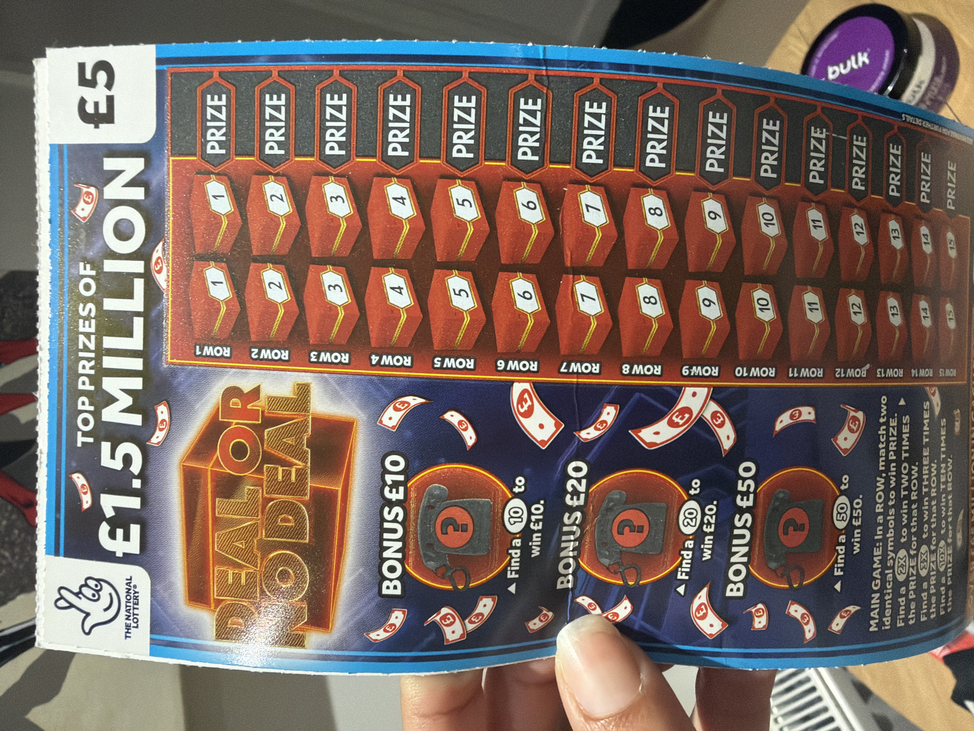 A slightly creased "Deal or No Deal" scratch card from The National Lottery is held by a person's left hand, with the thumb visible on the left side of the card. The card features a blue background with gold and red elements. The top section displays "TOP PRIZES OF £1.5 MILLION" and the ticket price of "£5". The logo for "The National Lottery" (a hand holding a ball) is in the upper left corner.
The main game section on the right side of the card consists of 15 rows, labeled "ROW 1" through "ROW 15". Each row contains two red box-like symbols with white numbers (1 to 15) and a "PRIZE" box to the right. The instructions for the main game, partially visible at the bottom, state: "MAIN GAME: In a ROW, match two identical symbols to win PRIZE. Find a 2X to win TWO TIMES the PRIZE for that ROW. Find a 3X to win THREE TIMES the PRIZE for that ROW. Find a 10X to win TEN TIMES the PRIZE for that ROW."
On the left side of the card are three bonus games: "BONUS £10", "BONUS £20", and "BONUS £50". Each bonus game features a grey rotary telephone icon with a red question mark in the center, surrounded by depictions of falling pound sterling notes. The instructions for these bonus games state: "Find a (10/20/50) to win £10/£20/£50."
In the lower-left background, a white, ribbed object, possibly a radiator or vent, is partially visible. In the lower-right background, a dark purple circular container with "bulk" written in white text is visible. The immediate background is blurred, suggesting an indoor setting.