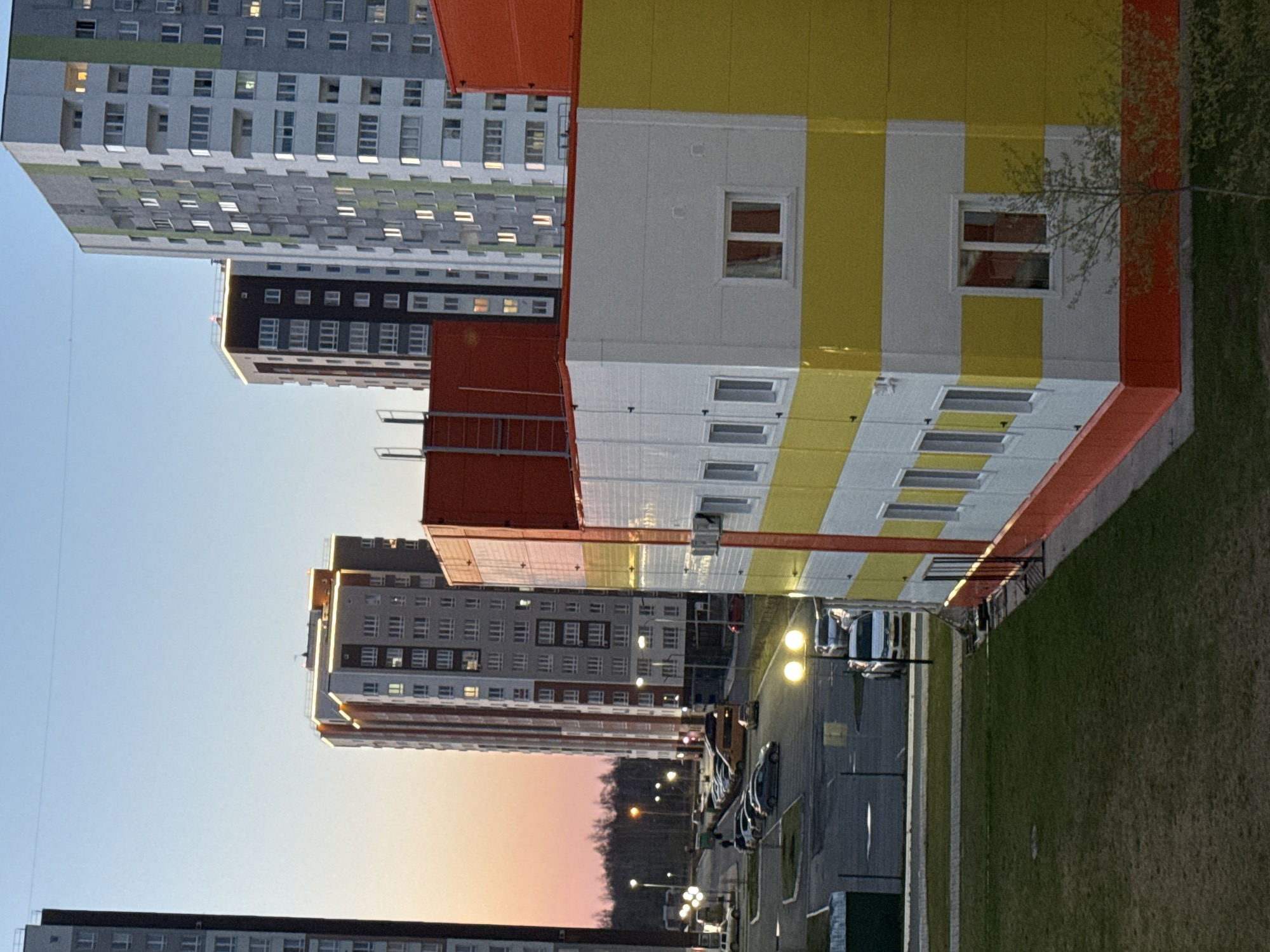 The image depicts a cityscape in Tobolsk, Russia, at dusk. Several high-rise apartment buildings dominate the scene, with colorful facades featuring orange, yellow, and white stripes. Rows of windows indicate residential occupancy. A street with parked cars and streetlights runs between the buildings and a green lawn. The sky transitions from blue to pink near the horizon.