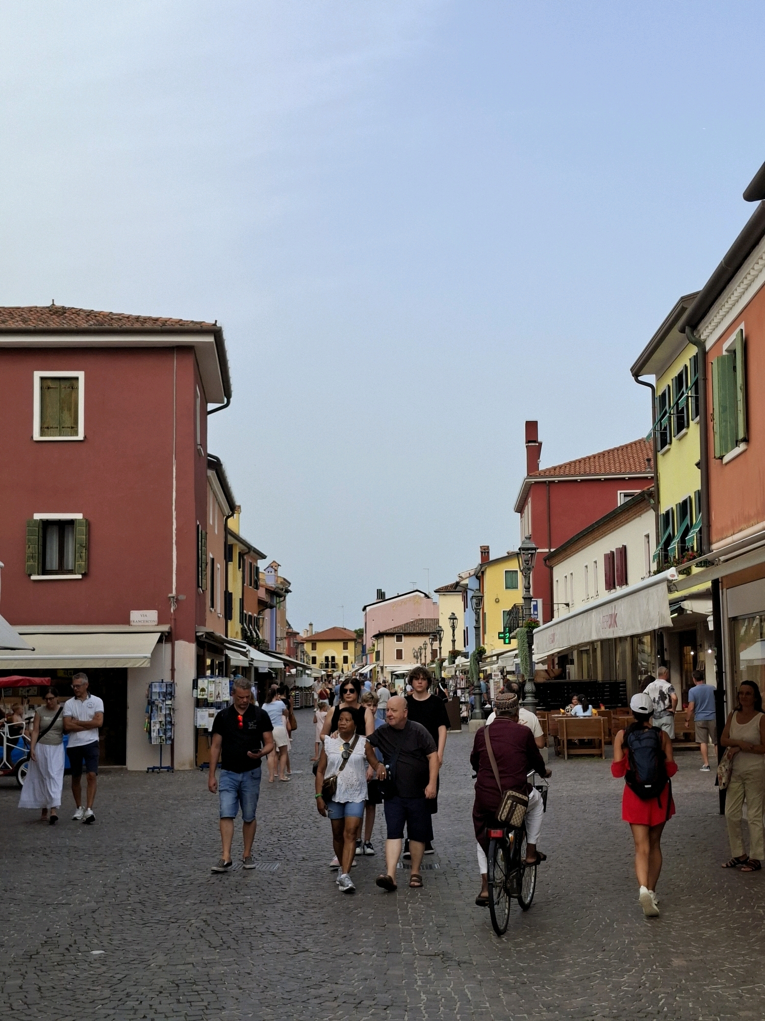 The image depicts a street scene in Caorle, Italy, likely a pedestrianized area with shops and restaurants lining both sides. The buildings are brightly colored, with predominantly red, yellow, and orange facades, featuring traditional shutters and awnings. The street is paved with cobblestones. Several people are walking along the street, some individually and some in small groups.
In the foreground, a man in a dark t-shirt and denim shorts is walking towards the camera, looking at his phone. To his right, a woman in a white top and denim shorts walks away from the camera, with a backpack. Further ahead, a man is riding a bicycle, also moving away from the camera.
On the left side of the street, a couple is walking towards the camera. The woman is wearing a white dress, and the man is in a white polo shirt and blue shorts. Other individuals and groups of people are visible further down the street, suggesting a lively atmosphere.
The sky is clear and light blue, indicating daytime, possibly late afternoon or early evening given the soft lighting. The overall impression is a vibrant and bustling European street scene.