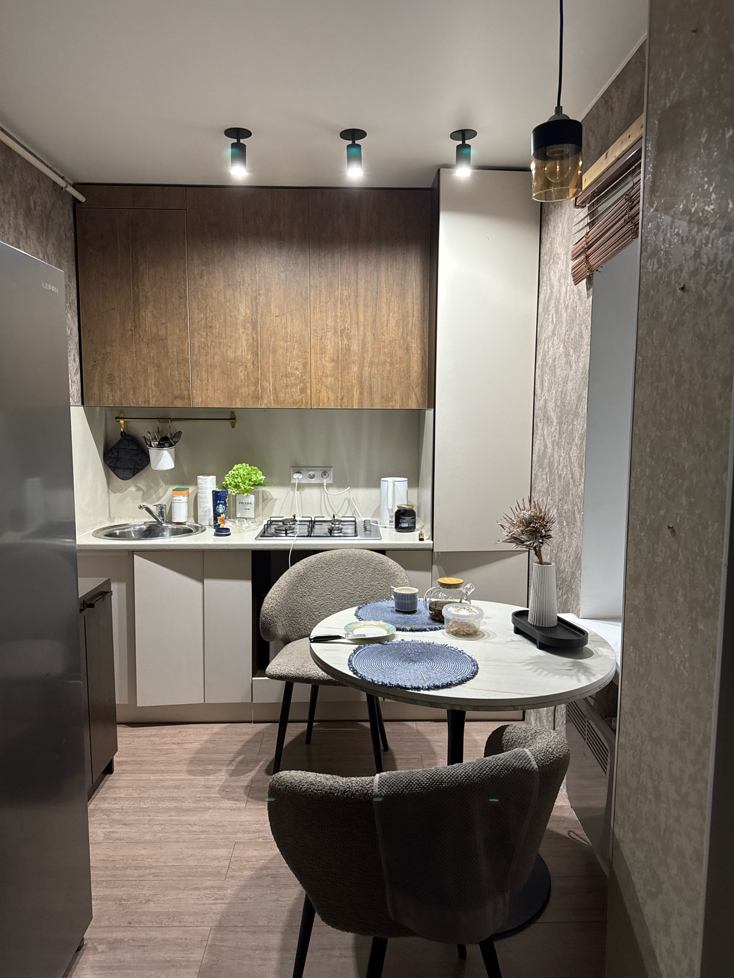 The image shows a compact kitchen with a dining area. On the left, a stainless steel refrigerator is partially visible. The kitchen counter features a sink, a stovetop, and various items including a plant, containers, and a coffee maker. Above the counter are wooden cabinets. A round dining table with two chairs is placed in the center of the room. The table is set with placemats, cups, and a small vase with dried flowers. The lighting in the kitchen includes ceiling spotlights and a hanging pendant light. The walls have a textured wallpaper. The floor is wood-like laminate. The scene appears to be an indoor residential kitchen. The location of Valday, Russia is not depicted in the image.