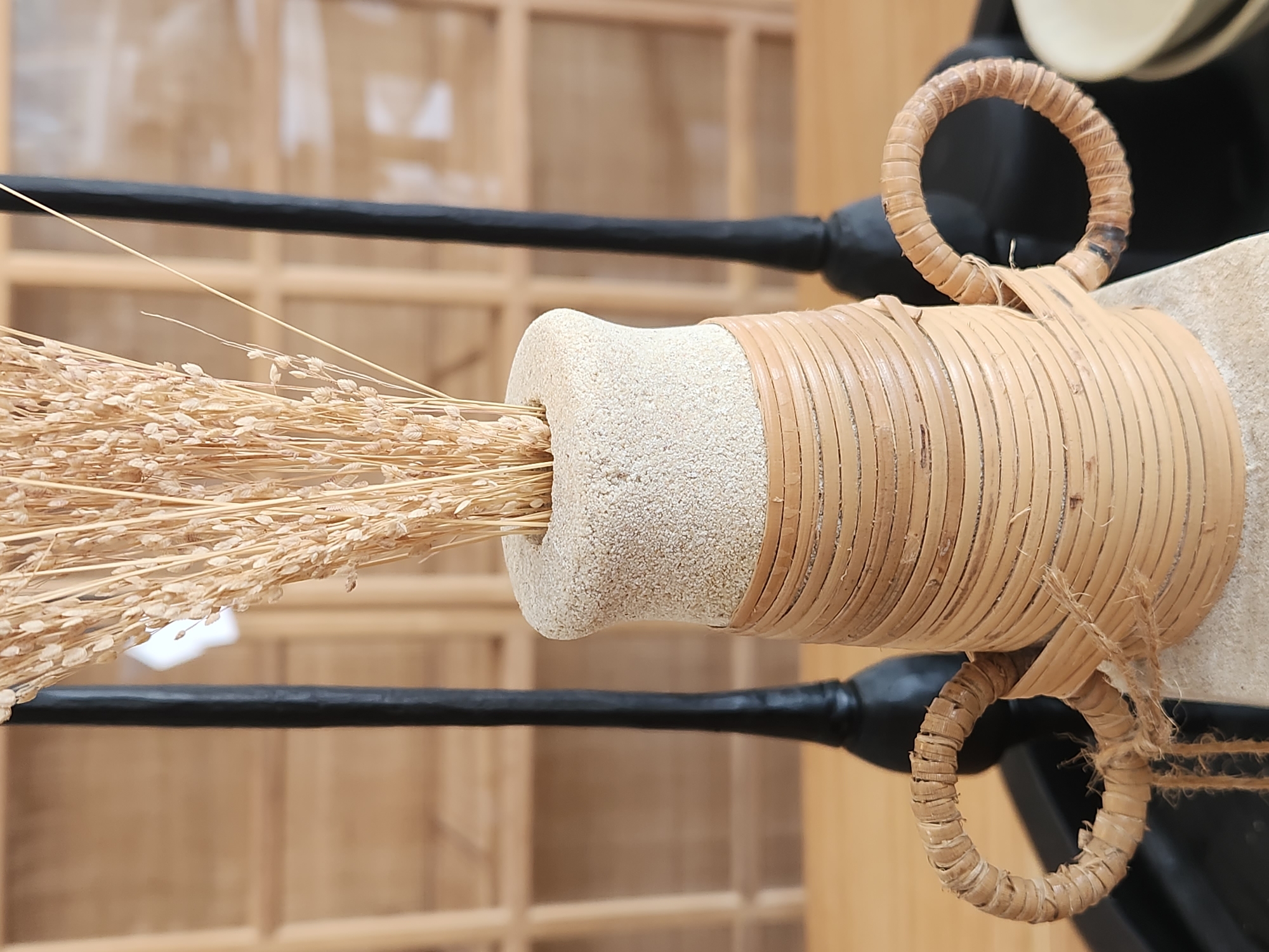 The image shows a close-up of a decorative vase filled with dried wheat stalks. The vase has a textured, sand-colored top and a woven rattan body with two looped handles. In the background, two tall, slender black candlesticks are visible, and behind them, a wooden shelving unit. The scene is set on a wooden table. The image does not contain any people or any identifiable landmarks, making it impossible to determine its location within Maitland, United States of America.