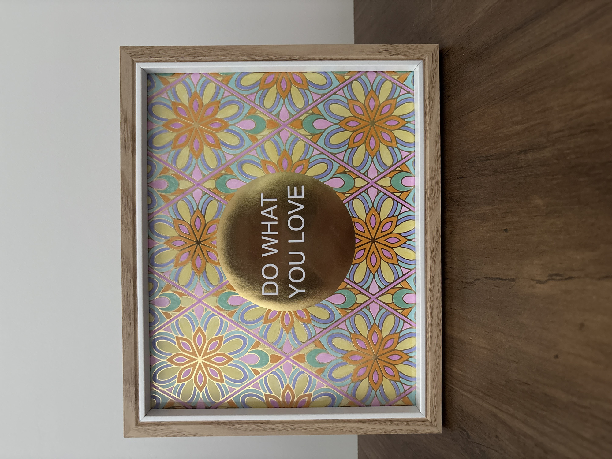 A framed picture with a pastel floral pattern and the text "DO WHAT YOU LOVE" in gold is displayed on a wooden surface. The frame is made of light-colored wood with a white inner border. The background is a plain white wall. The image does not provide any information about the city of Liberec, Czechia.