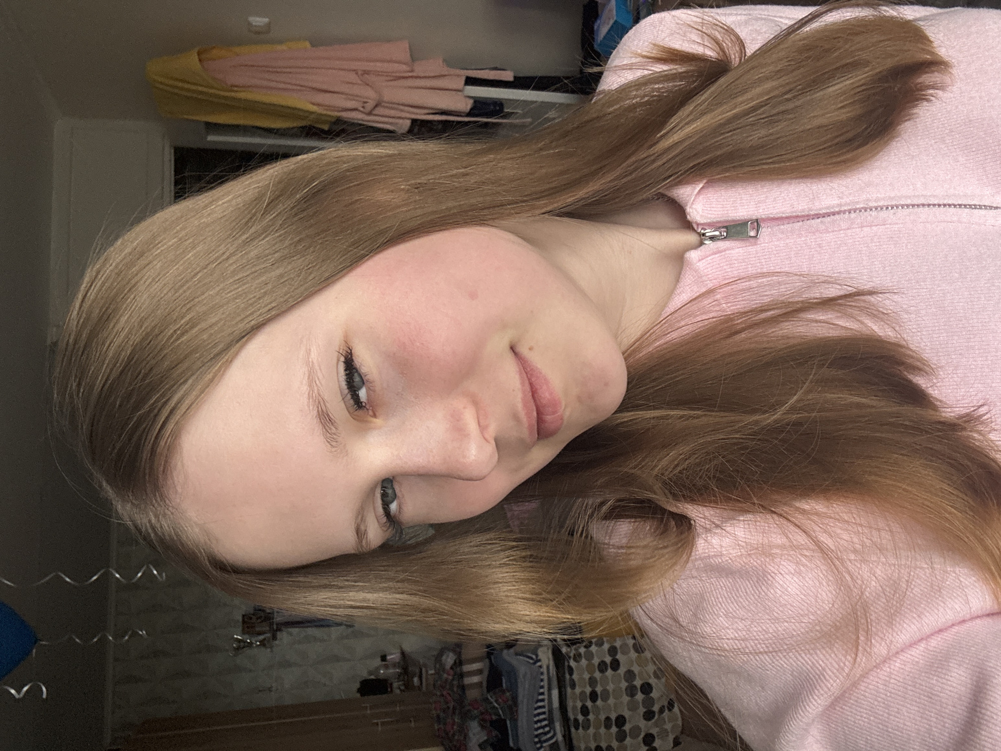 A portrait of a fair-skinned young woman with light brown hair, wearing a pink zippered sweatshirt. The background appears to be an indoor setting, possibly a bedroom, with glimpses of furniture, clothing, and decorations. The city of Chaikovsky, Russia, is not apparent in the image.